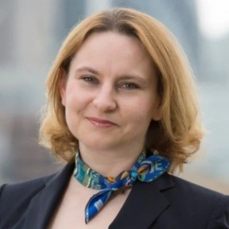 A woman with shoulder-length blonde hair, wearing a dark blazer and a blue patterned scarf, standing outdoors with a blurred cityscape background.