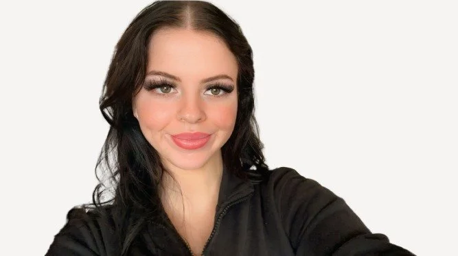 Young woman with long dark hair, makeup, and a black jacket taking a selfie against a white background.