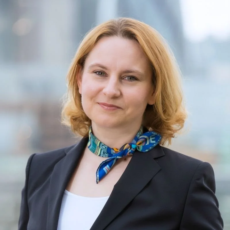 A woman with shoulder-length blonde hair, wearing a dark blazer and a blue patterned scarf, standing outdoors with a blurred cityscape background.