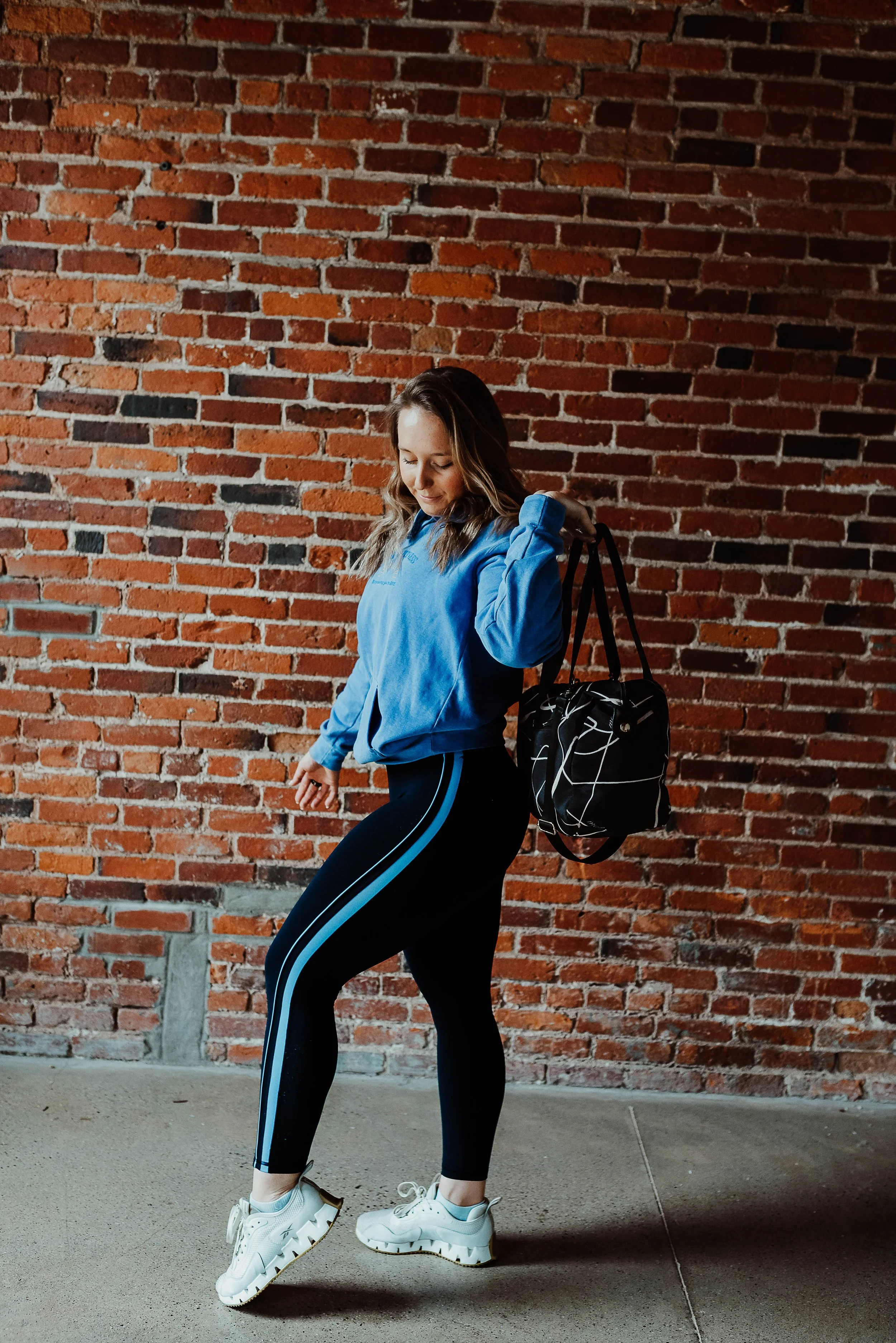 A woman wearing a blue hoodie and black athletic leggings with blue stripes, white sneakers, and a black patterned duffle bag over her shoulder, standing against a red brick wall, looking down and adjusting her sleeve.