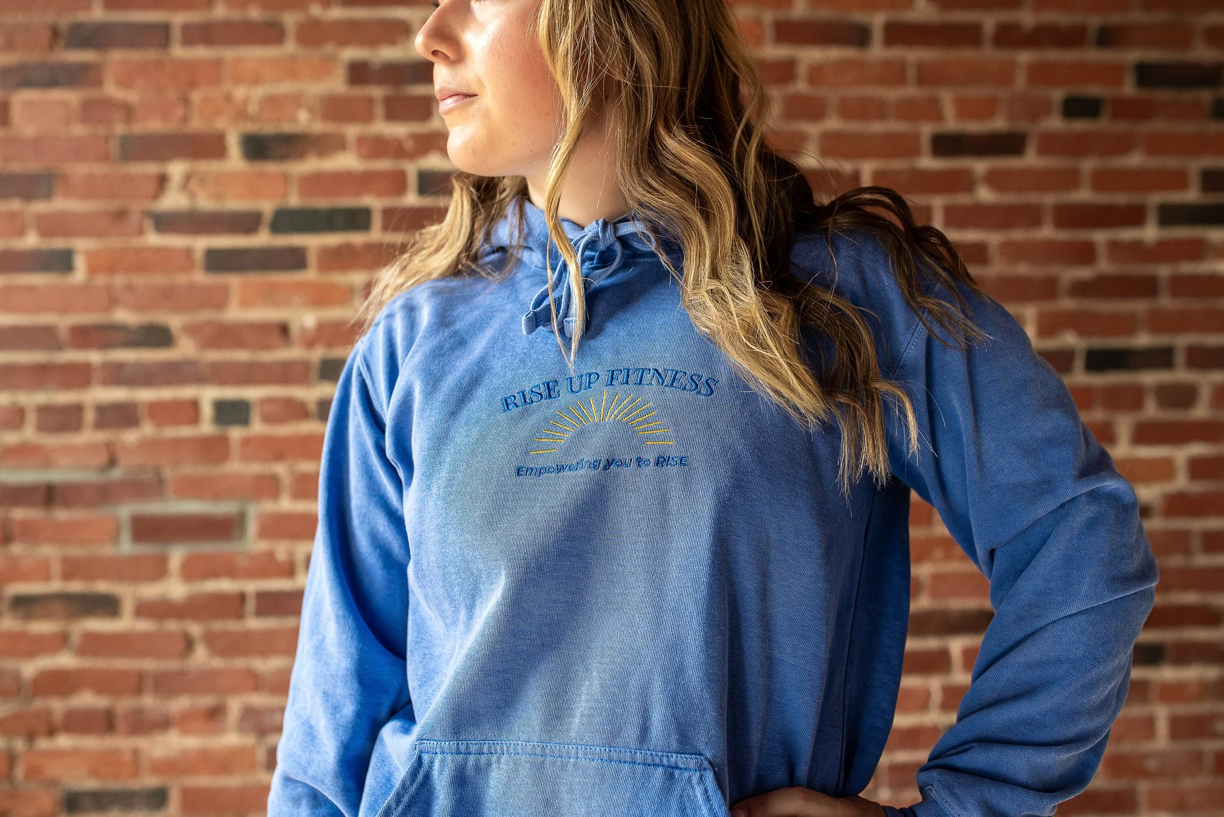 A woman with wavy blonde hair wearing a blue hoodie with the words "Rise Up Fitness" and a sun design, standing in front of a red brick wall.