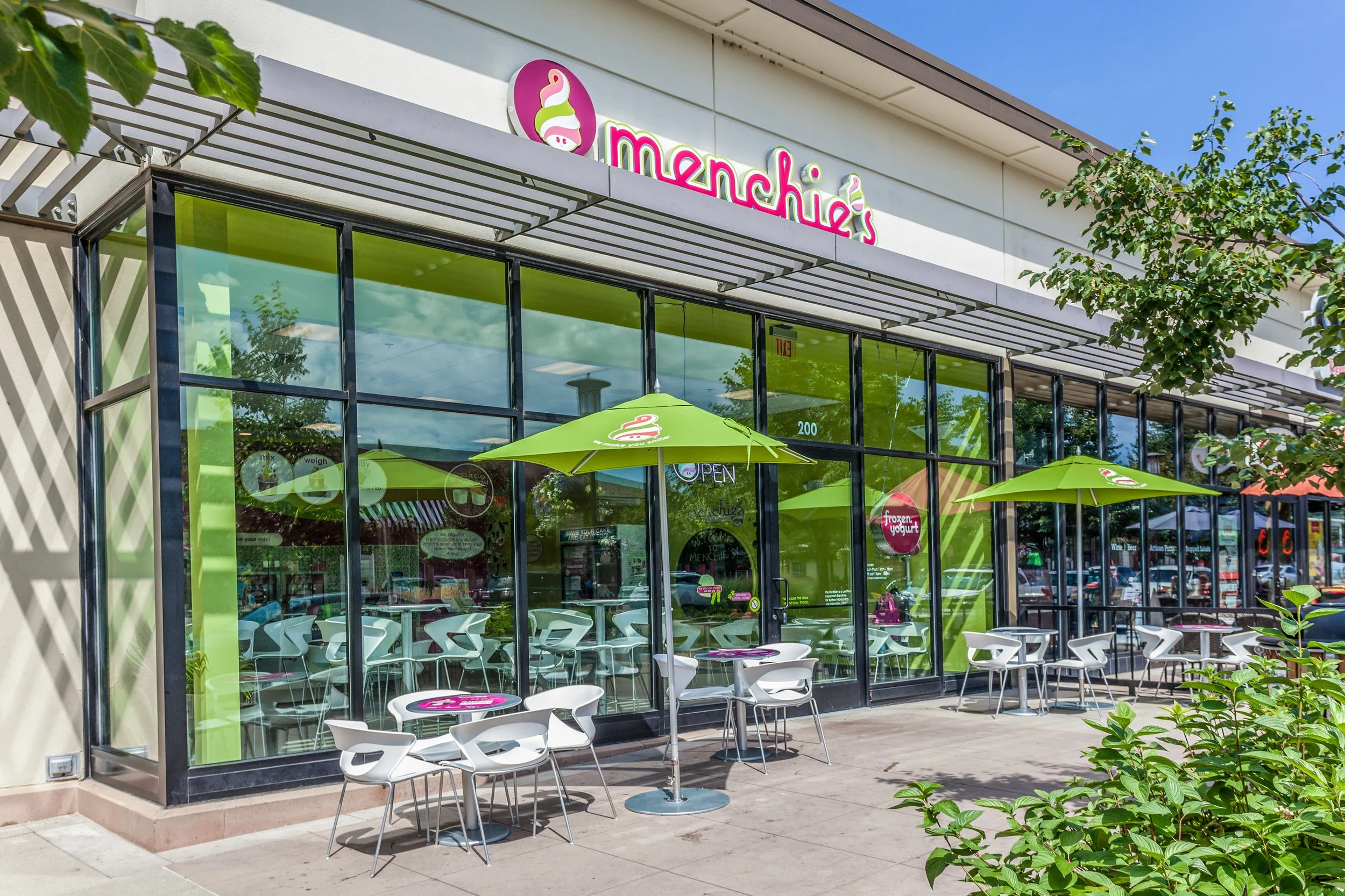 Exterior view of Menchie's frozen yogurt shop with large glass windows, outdoor seating with white tables and chairs, and green umbrellas. The shop's sign features a pink and green logo and the name Menchie's in pink letters.