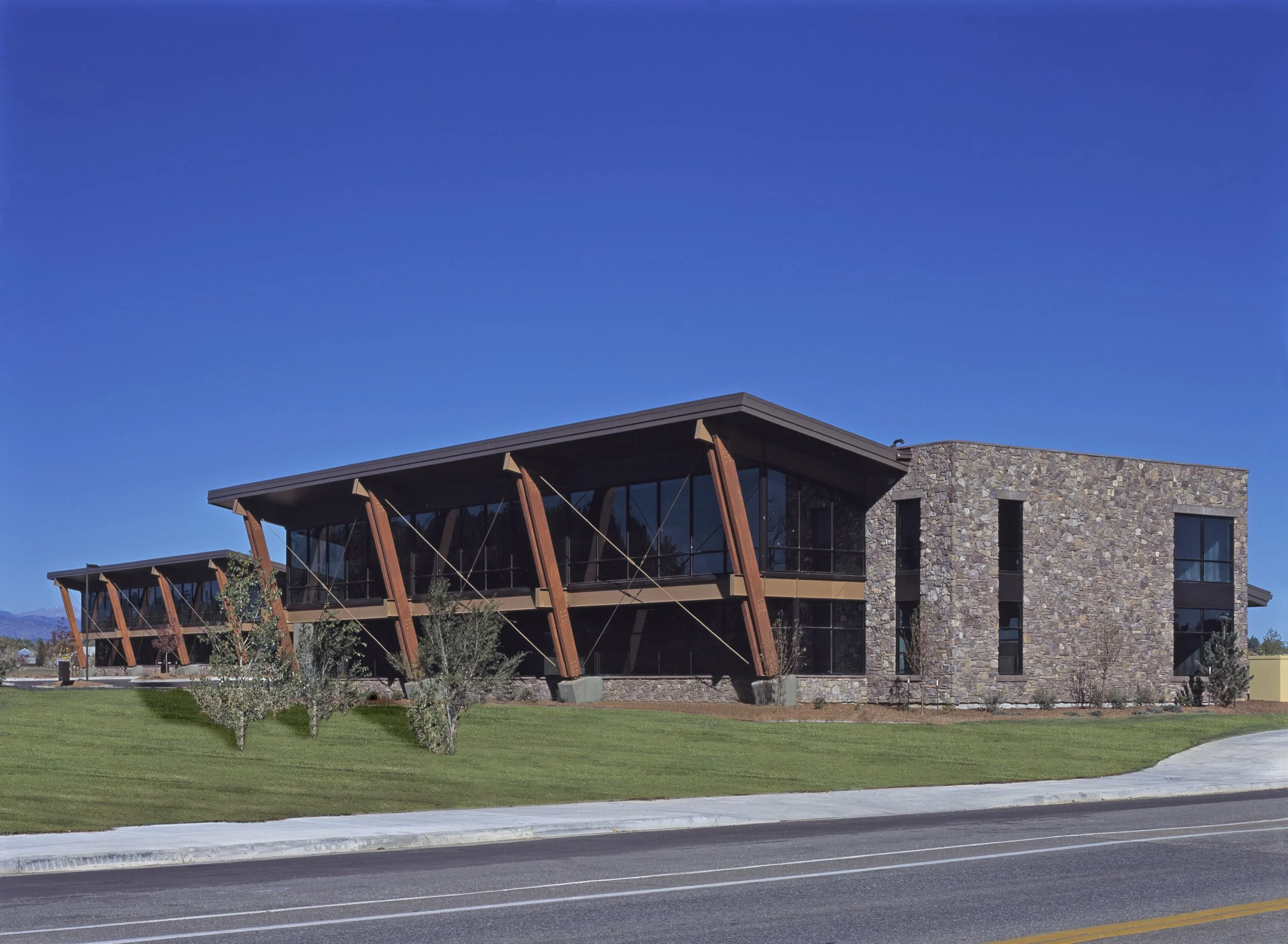 Modern building with stone and glass facade, large overhangs with wooden supports, trimmed in brown, surrounded by manicured grass and small trees, under a clear blue sky.