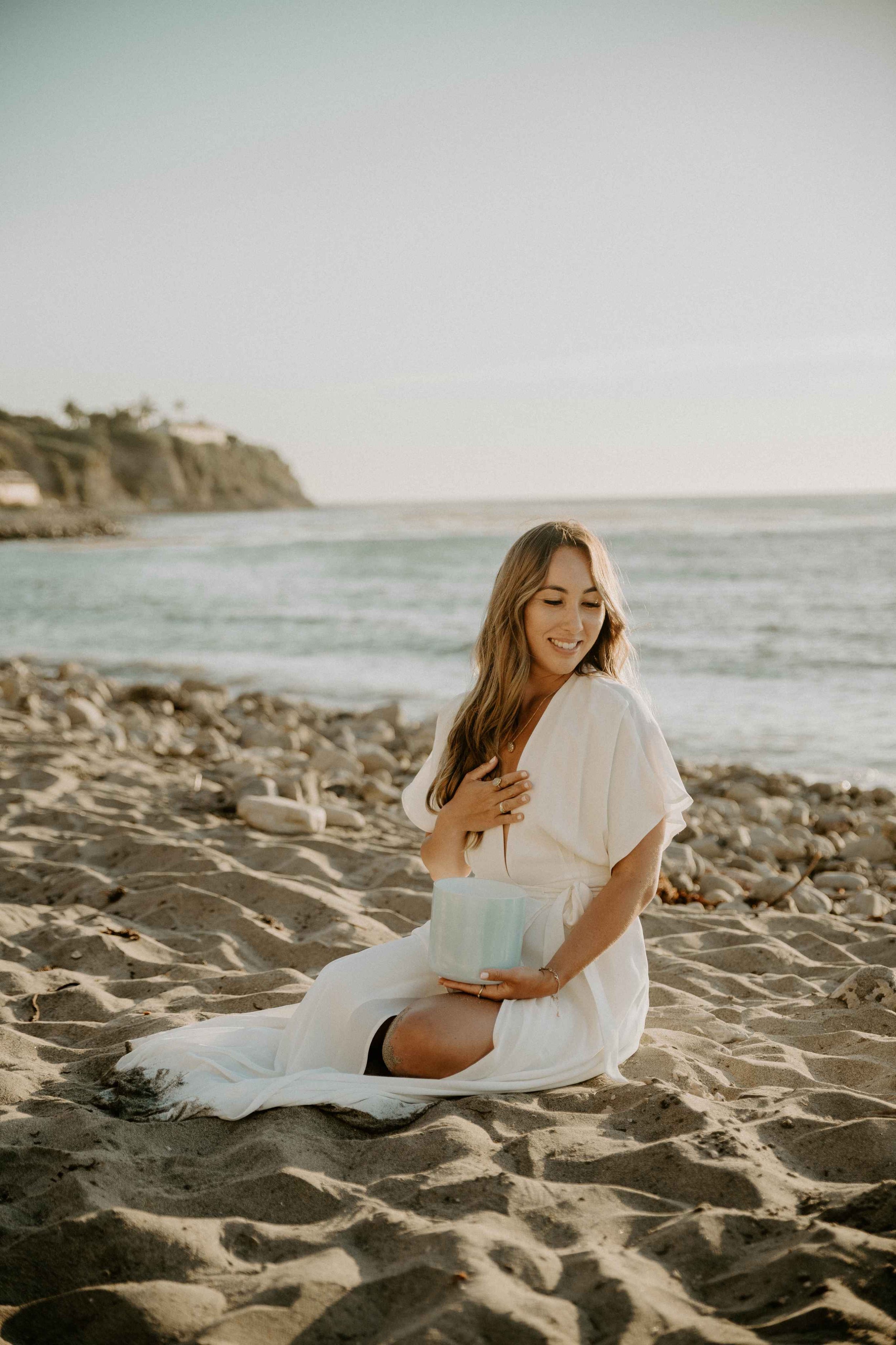 OHTP founder Sophie Pyne sitting on the sand at a beach in a white dress holding a sound healing bowl