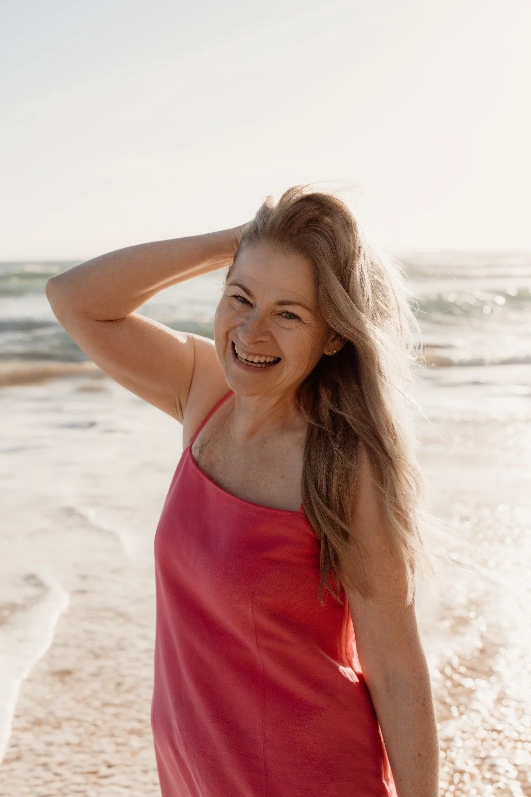 Carole Leigh in a bright pink dress by the ocean with her hand in her hair