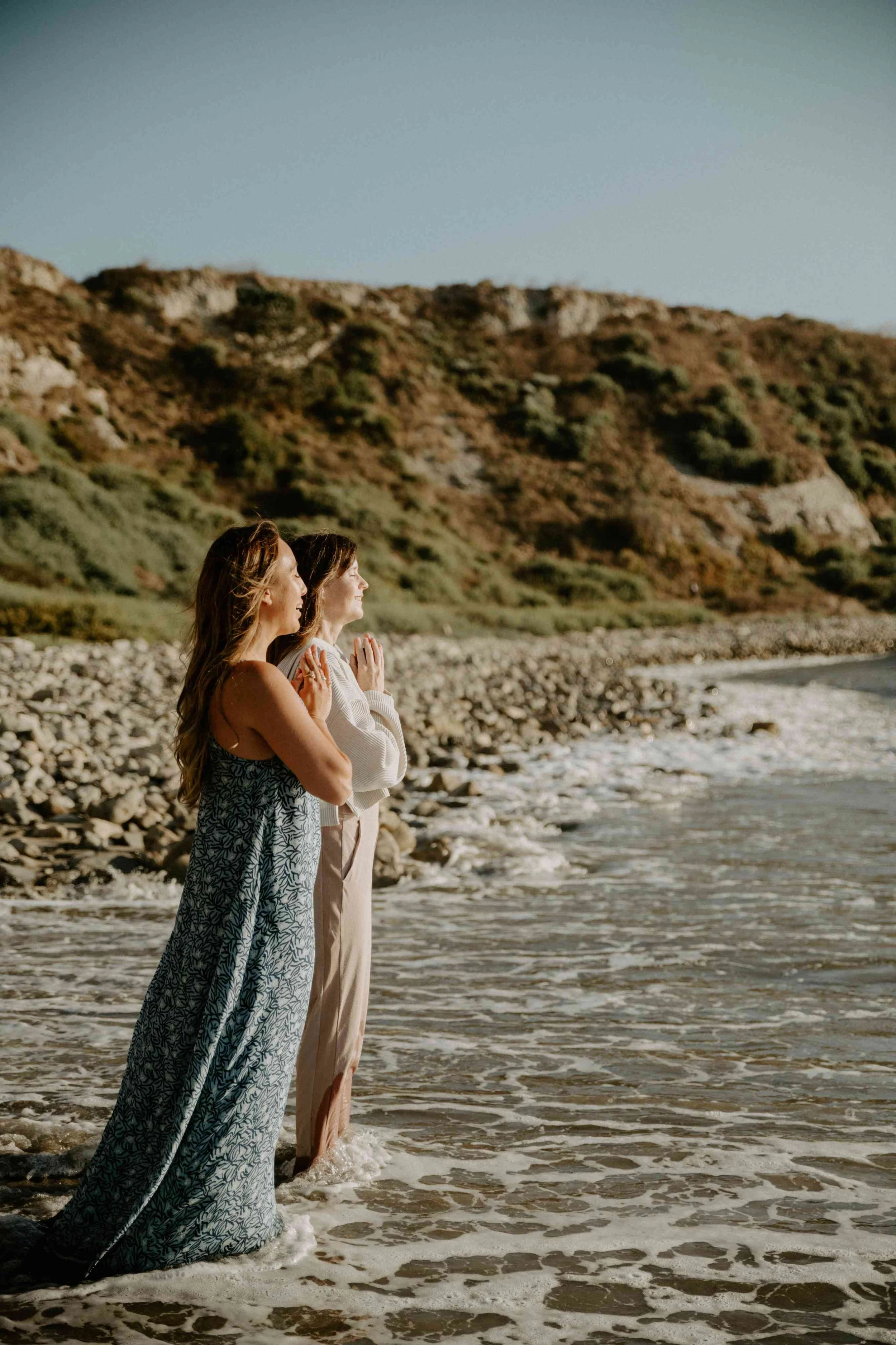 Two women standing in the ocean on the shore at sunset with rocky cliffs in the background