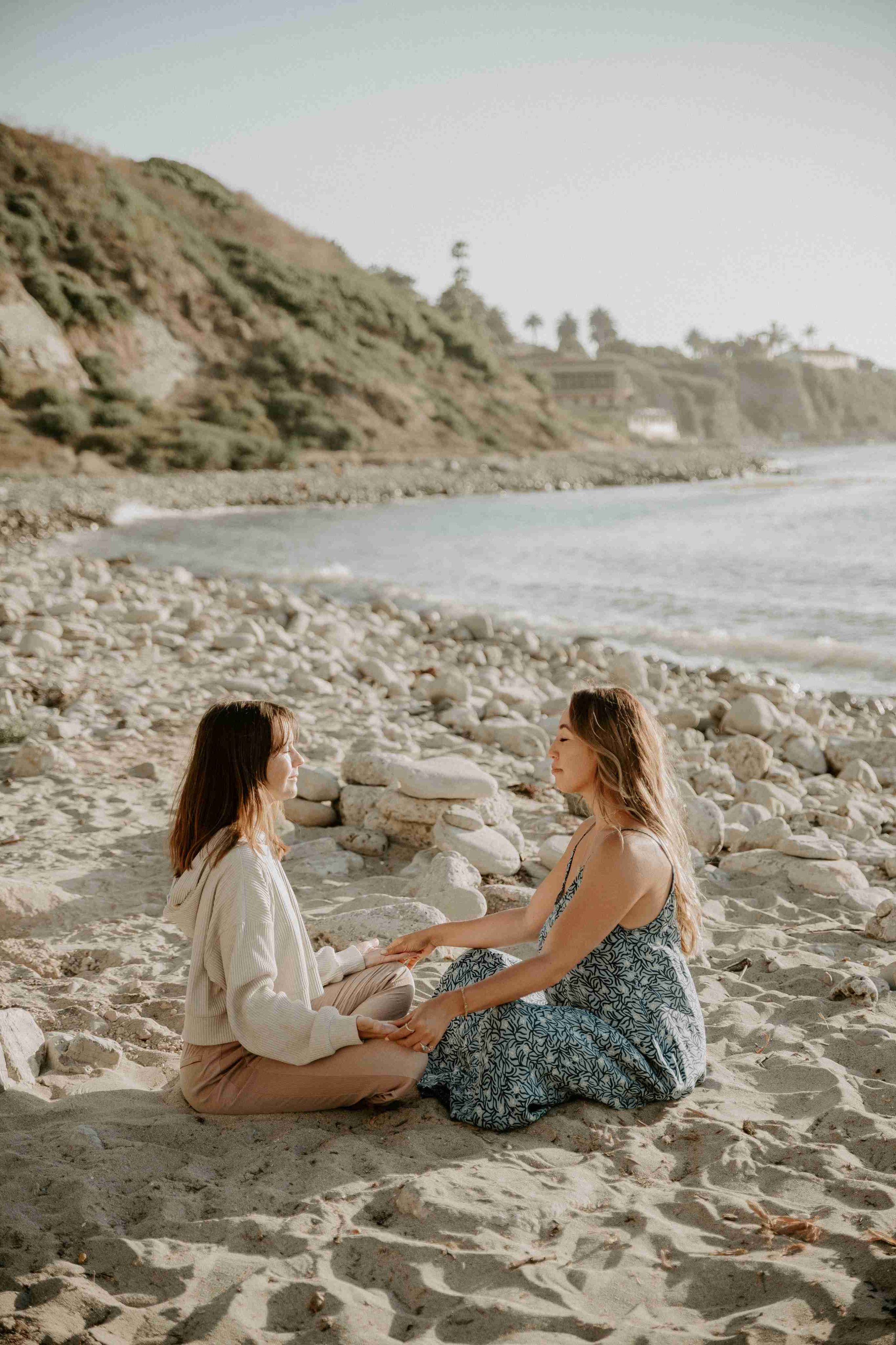 Two women sitting in ceremony on the beach by the ocean, meditating and holding hands