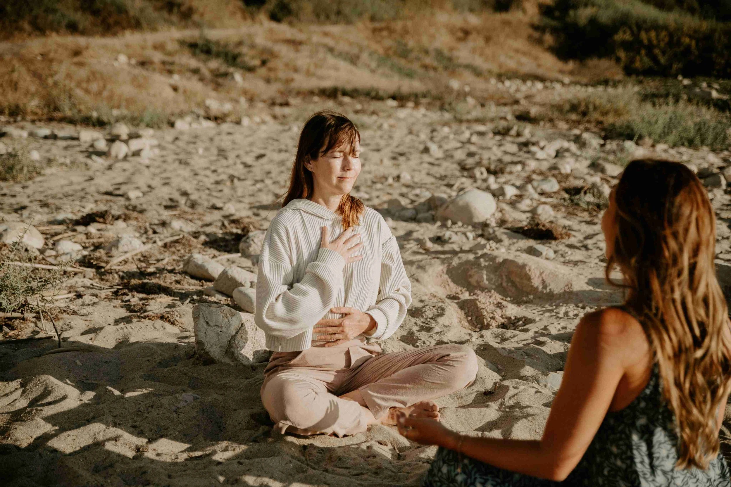 Two women meditating on a sandy rocky beach at sunset