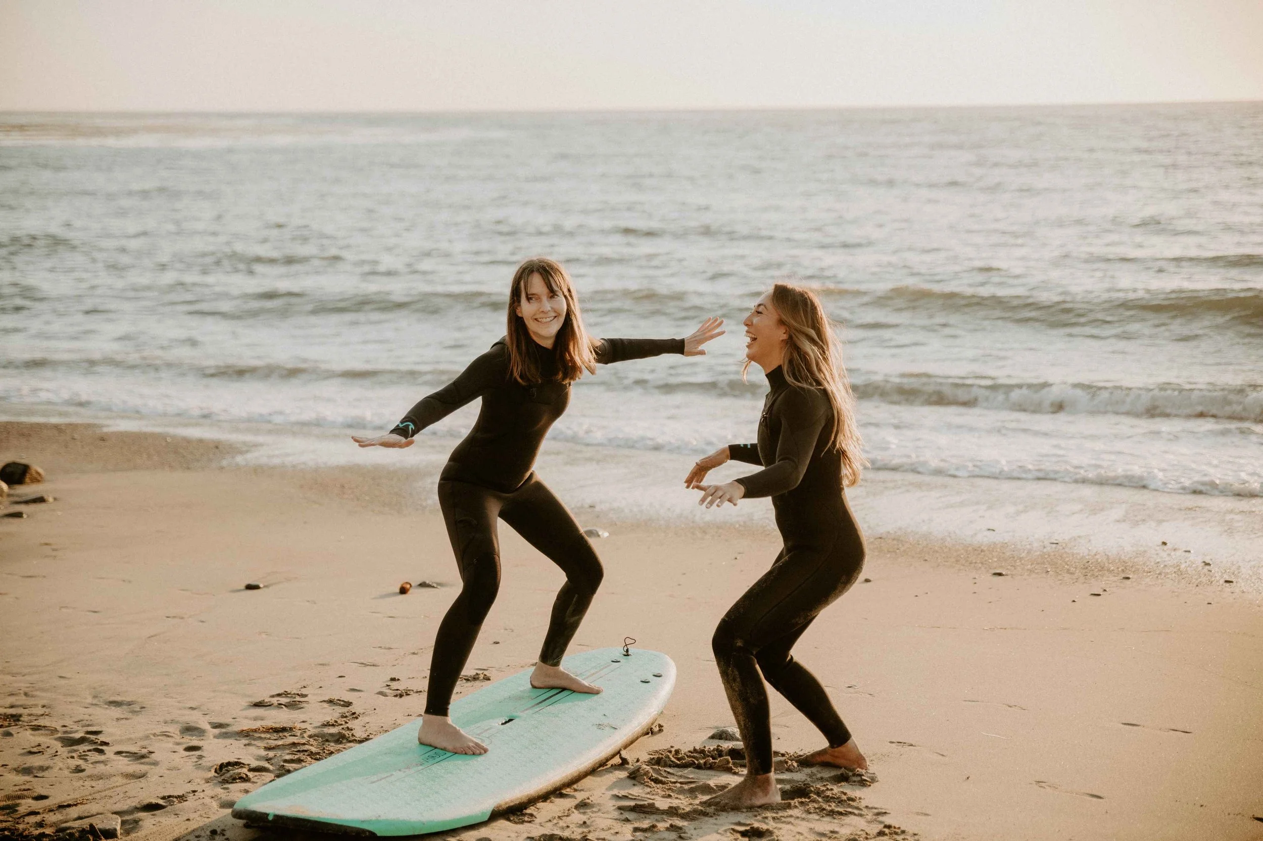 Two women at the beach getting ready to surf in wetsuits laughing and one is standing on a surfboard