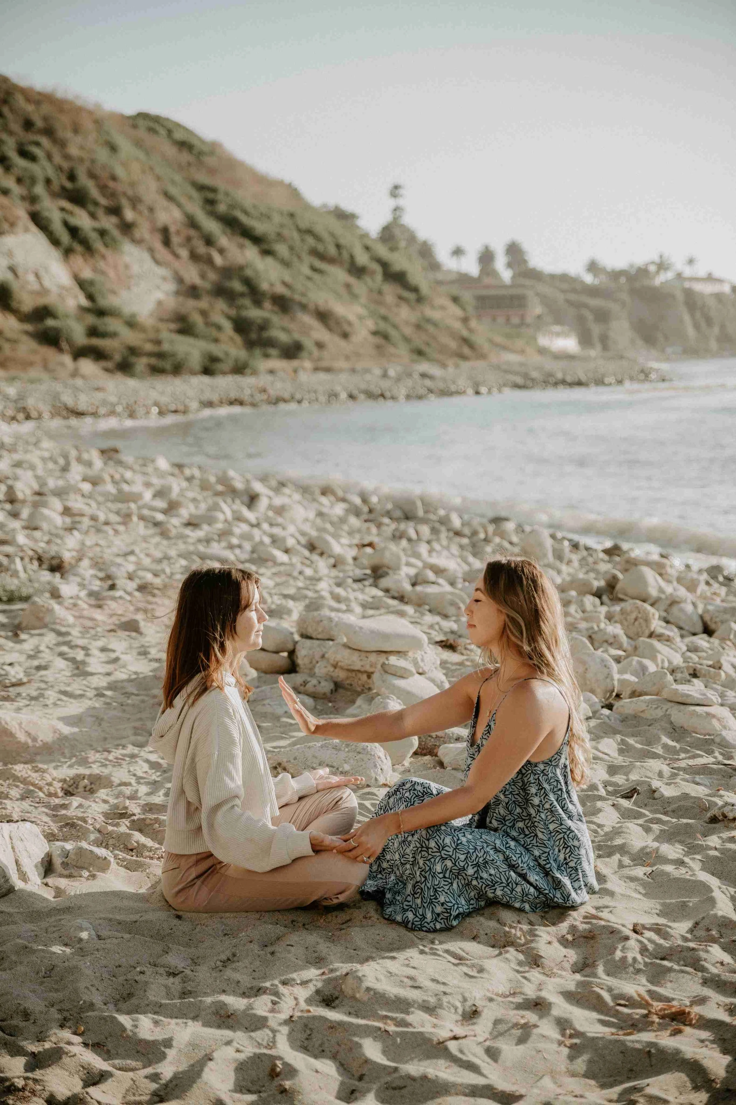 Two women sitting in the sand by the ocean in ceremony, holding hands and meditating with their eyes closed