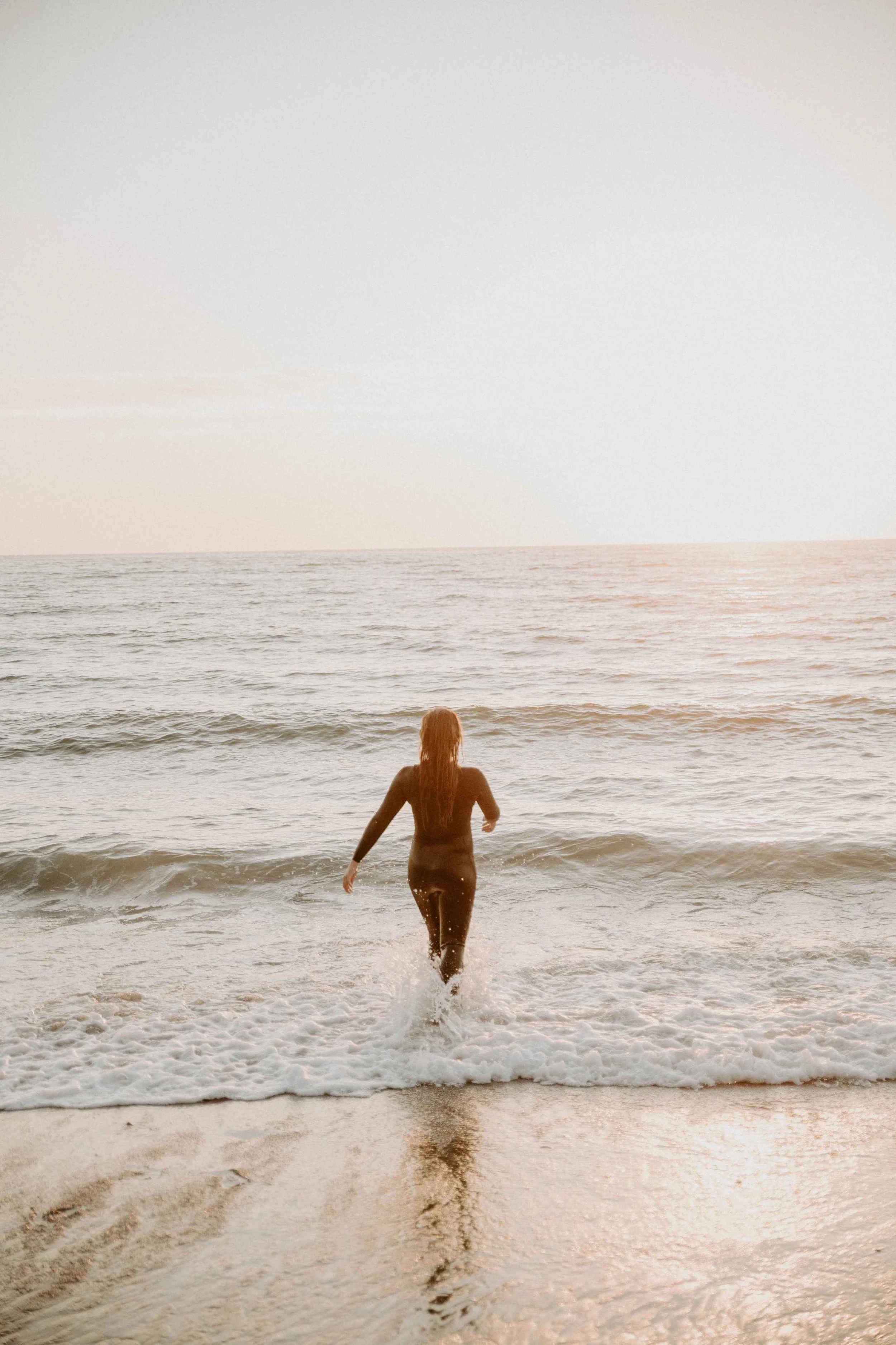 A woman walking into the ocean waves wearing a wetsuit at sunset