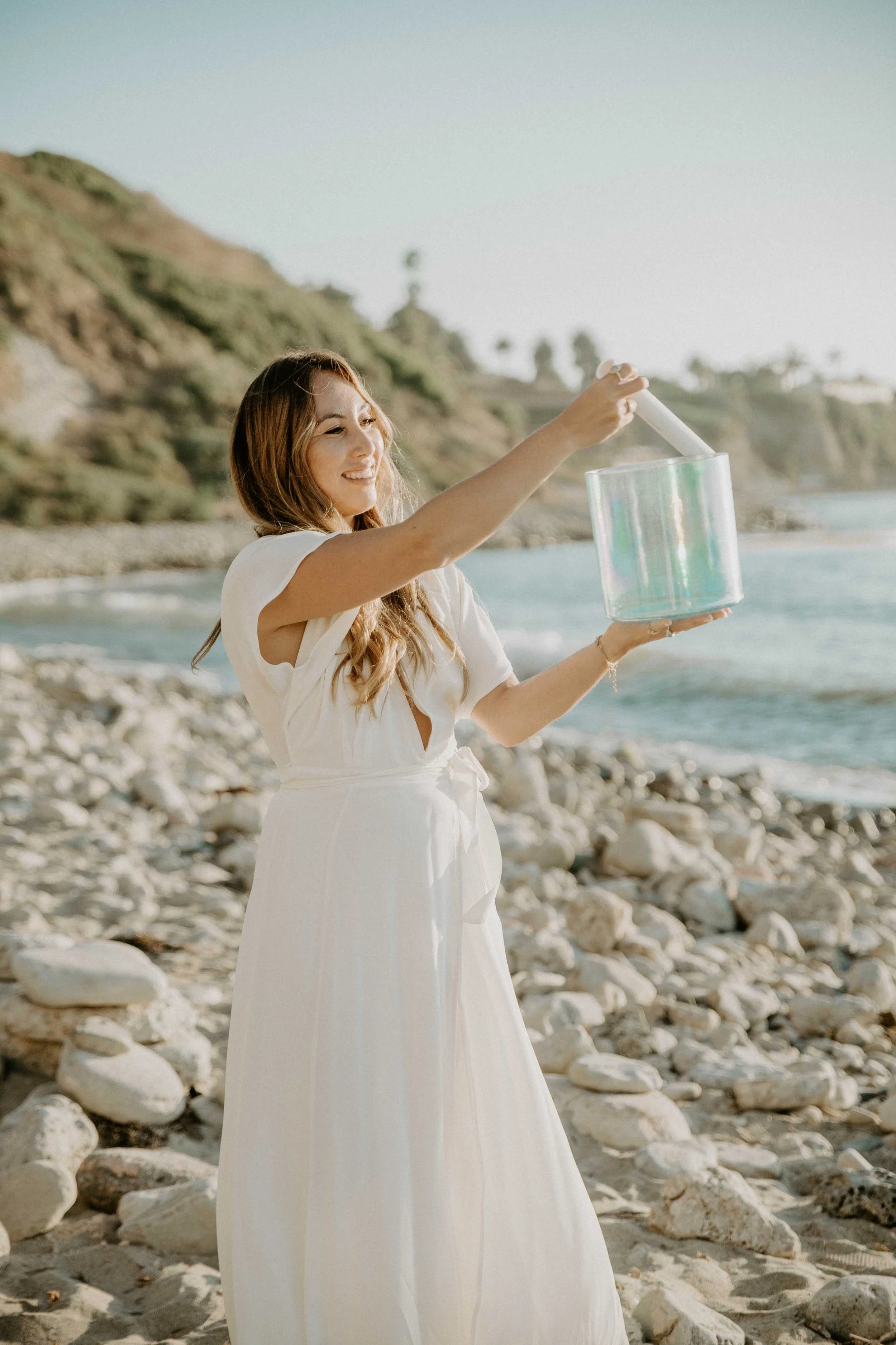Sophie Pyne doing sound healing with a white bowl by the ocean in a long white dress