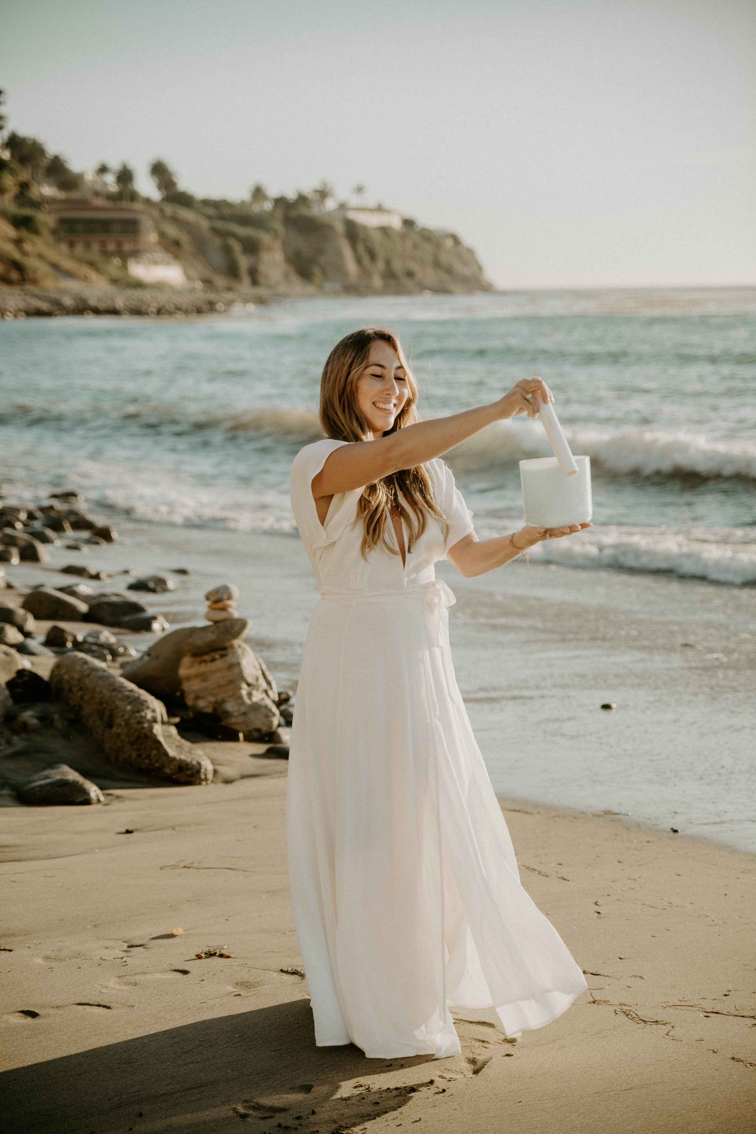 Sophie Pyne standing on the sand by the ocean in a long white dress with a white sound healing bowl