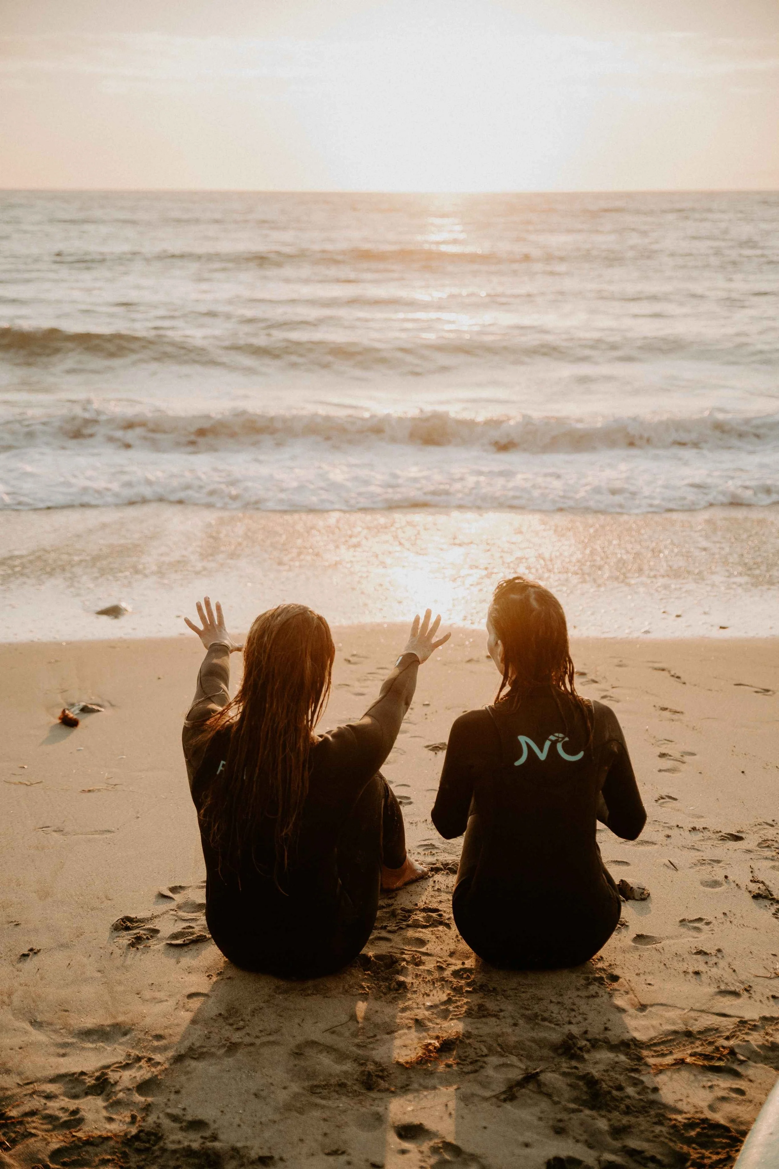 Two women sitting by the ocean on the sand at sunset wearing wetsuits and pointing to the water