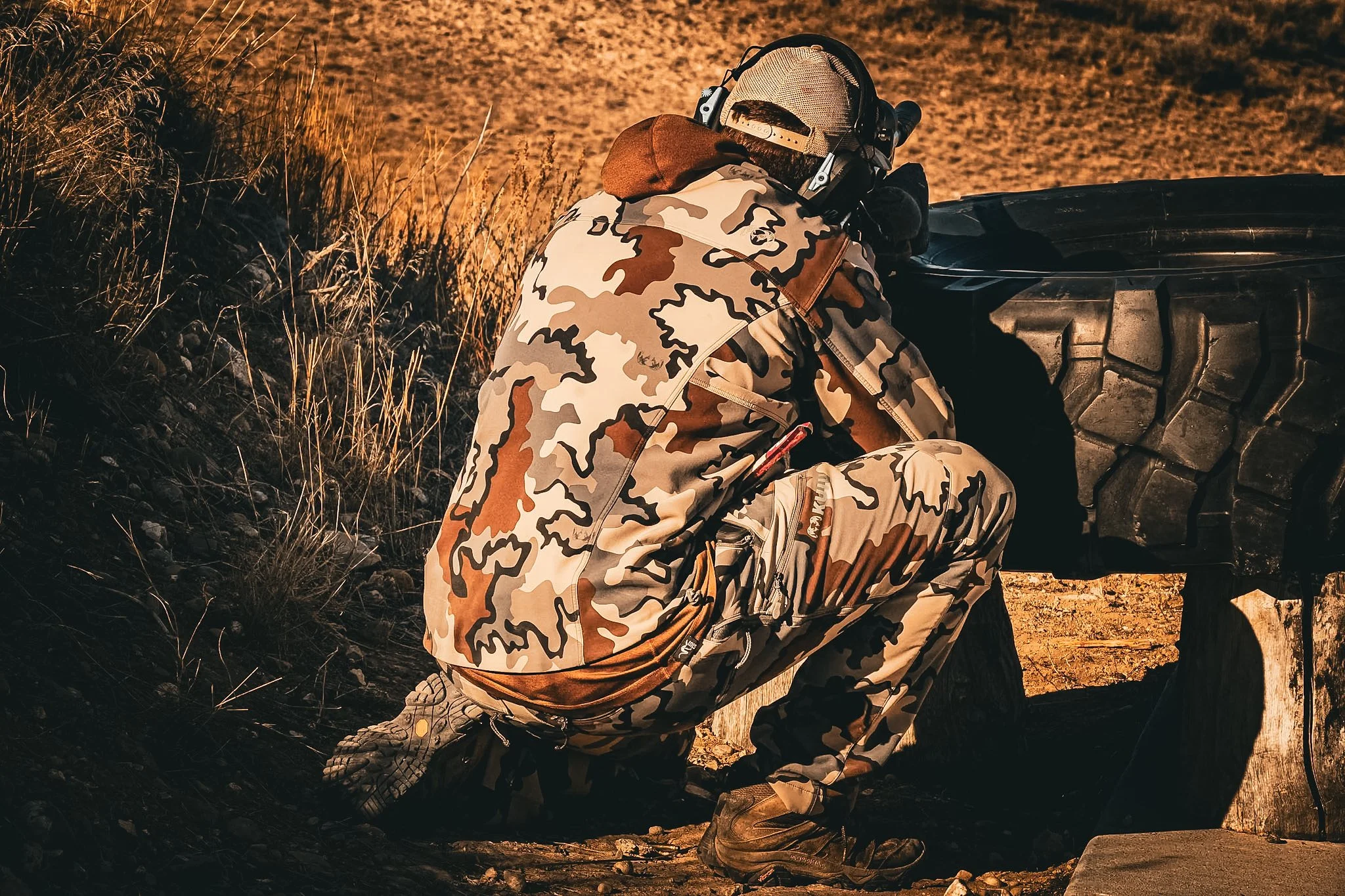 A person wearing camouflage clothing, kneeling down and inspecting the wheel of a large off-road tire, outdoors near a tree.