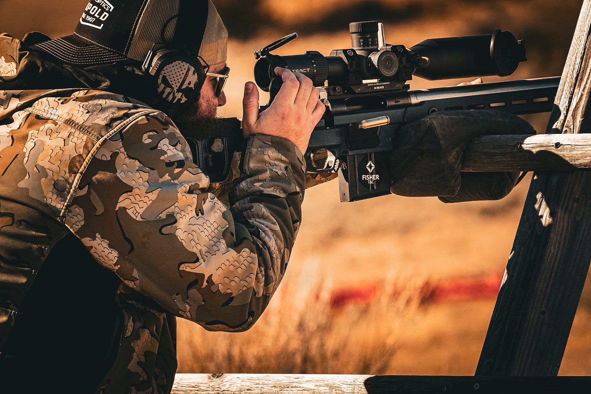 A man in camouflage clothing aiming a sniper rifle with a scope, leaning against a wooden structure outdoors during sunset.