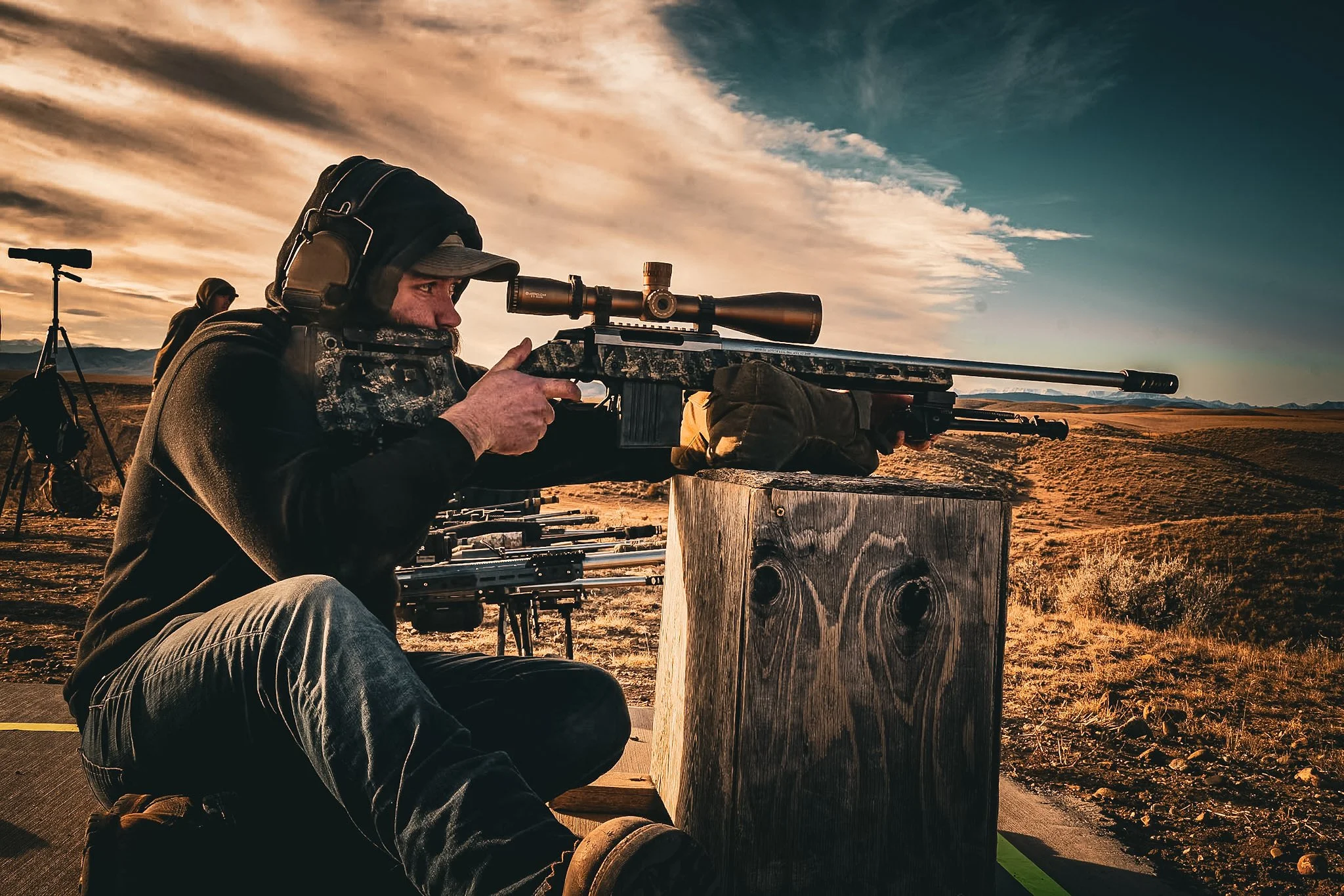 A man aiming with a scoped rifle during sunset in an open landscape.