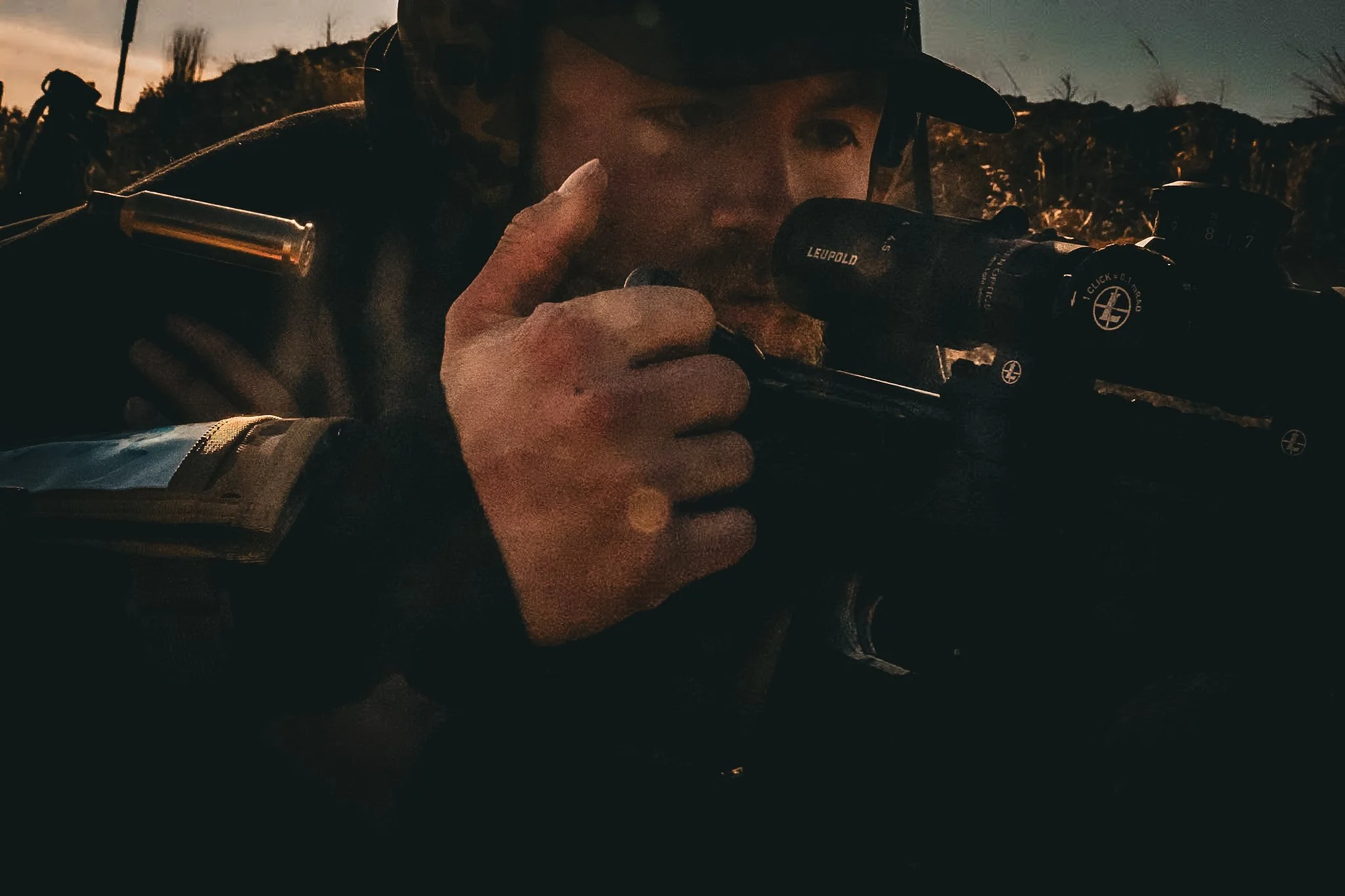 A woman in tactical gear wearing a helmet, aiming a sniper rifle with a scope during sunset or twilight.
