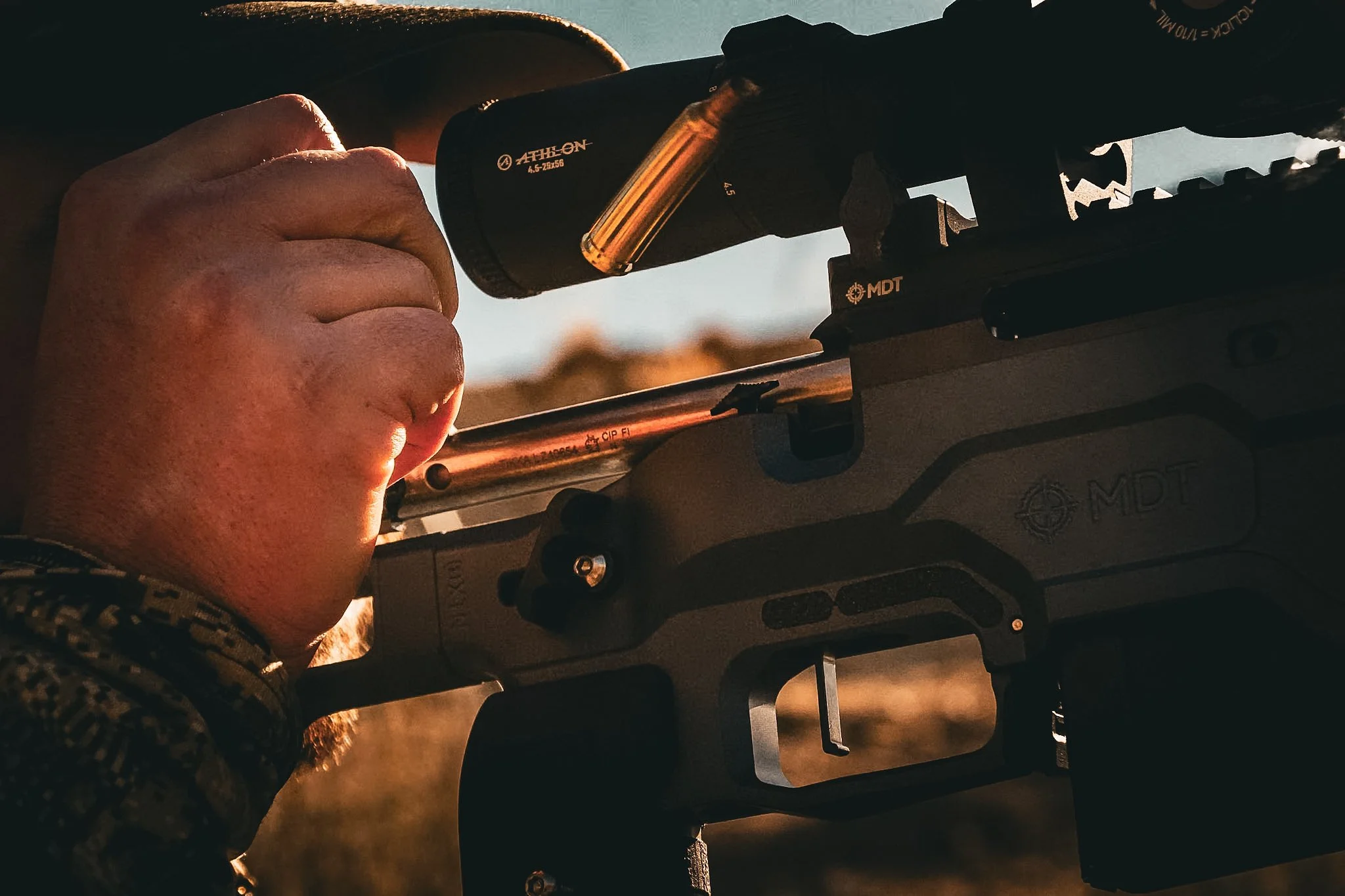 A person aiming a rifle outdoors at sunset, using a scope to view the target.