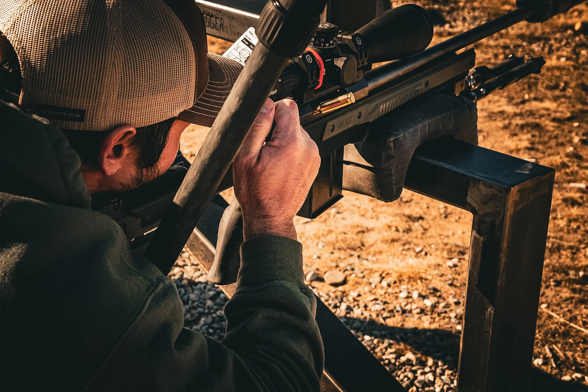A man wearing a beige cap and dark hoodie aiming a scoped rifle outdoors on a sunny day.
