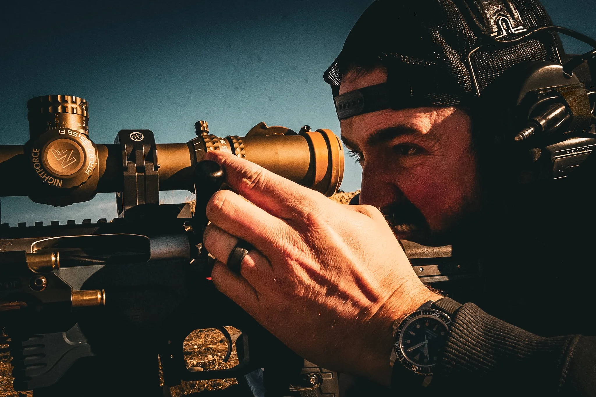 A man with a beard and wearing a black cap, aiming a sniper rifle with a golden scope, outdoors during daytime.