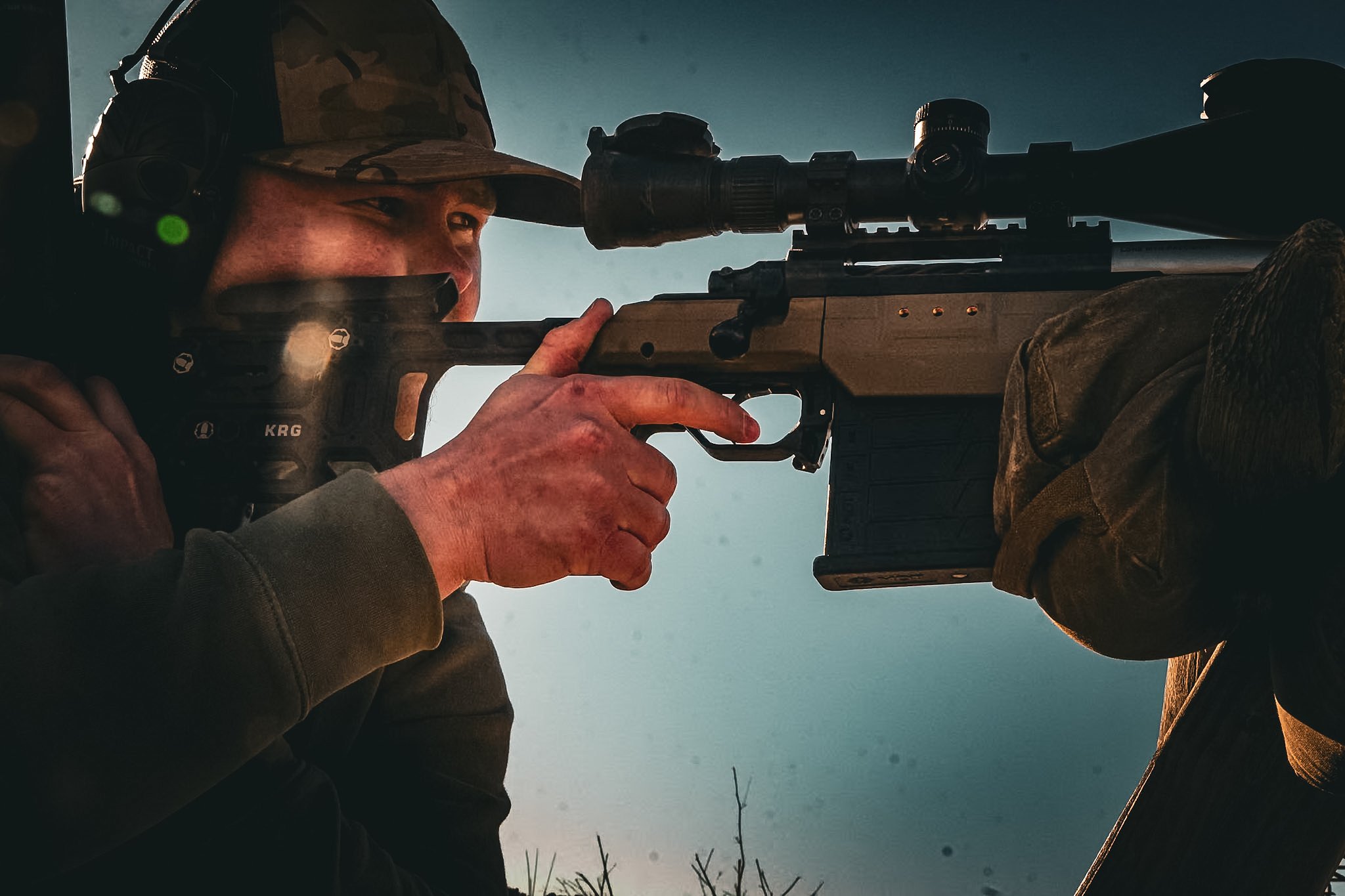 A person aiming a sniper rifle outdoors, wearing a camouflage hat and large headset for protection.