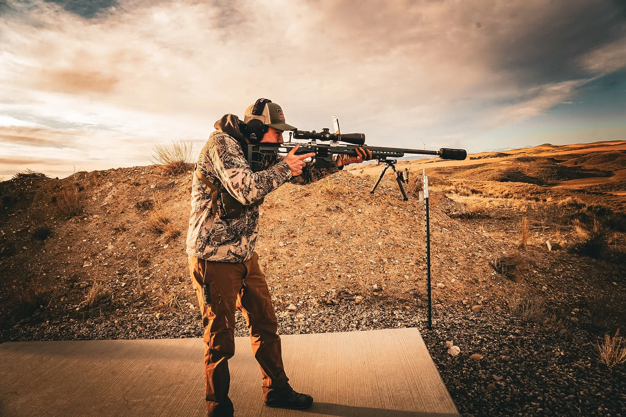 A person aiming a scoped rifle on a shooting range outdoors during sunset, with rolling hills in the background.