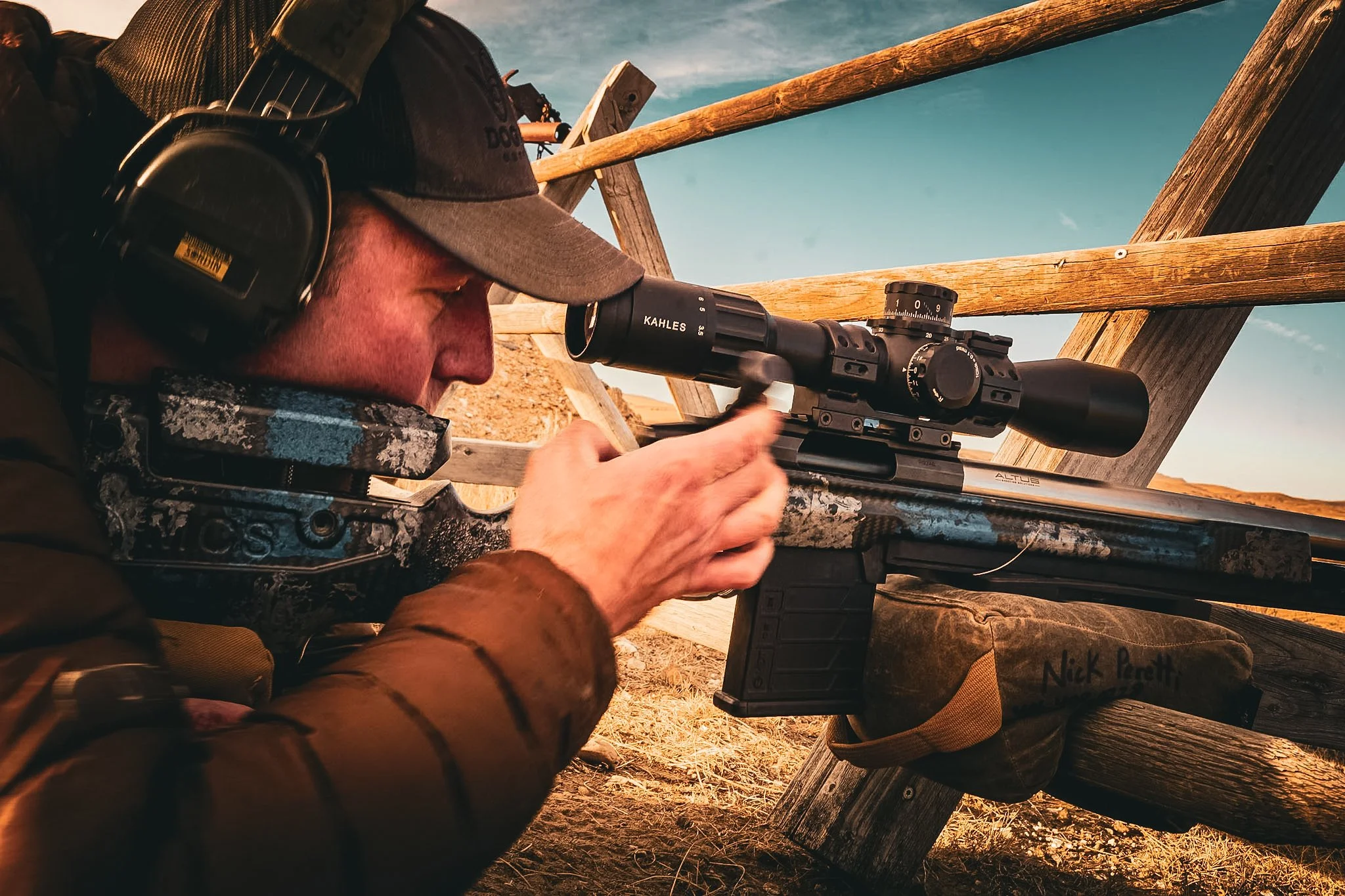 A man aiming a rifle through a scoped sight at an outdoor shooting range, wearing ear protection and a cap, with wooden barriers and a dirt ground in the background.