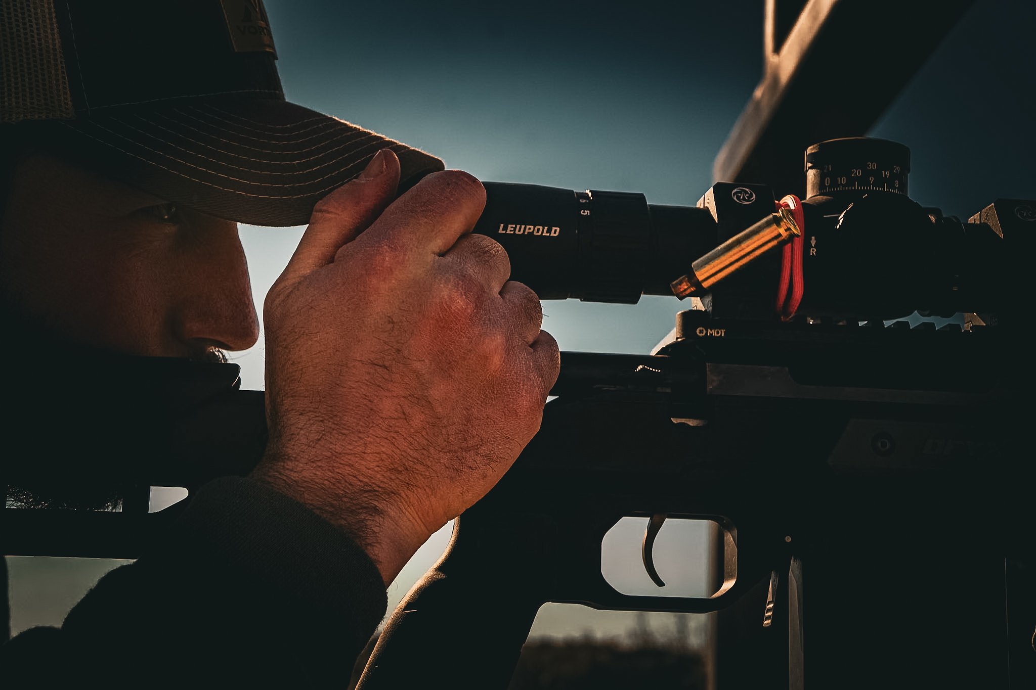 Person looking through a rifle scope outdoors, wearing a cap.