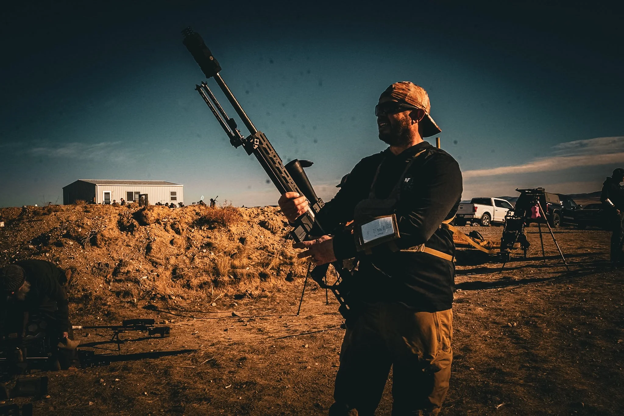 A person holding a large firearm in an outdoor setting during sunset, with several trucks and other people in the background.