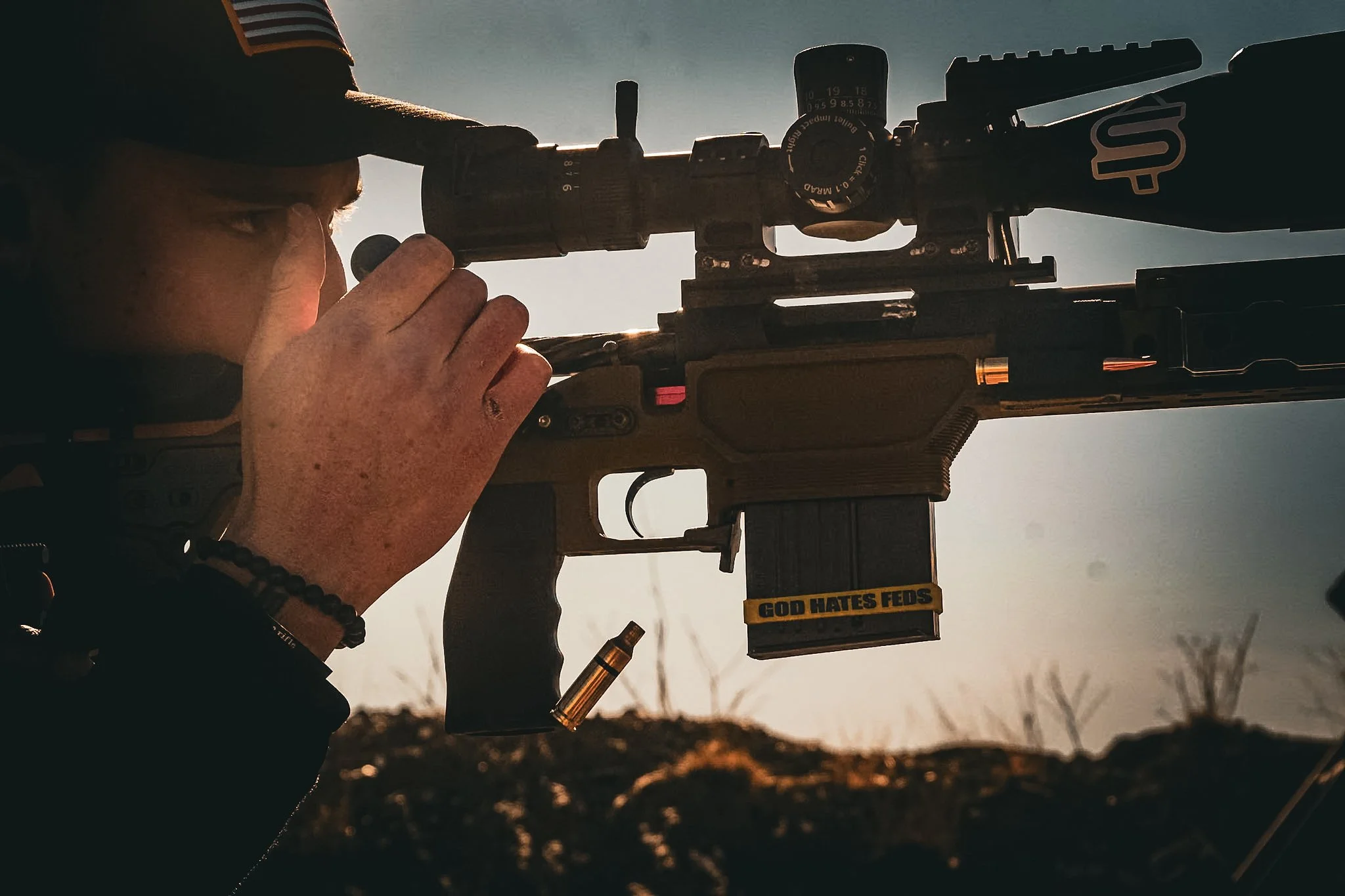 A person aiming a rifle with a scope, outdoors during sunset, with a sticker on the magazine that reads 'GOD HATES FEDS'.