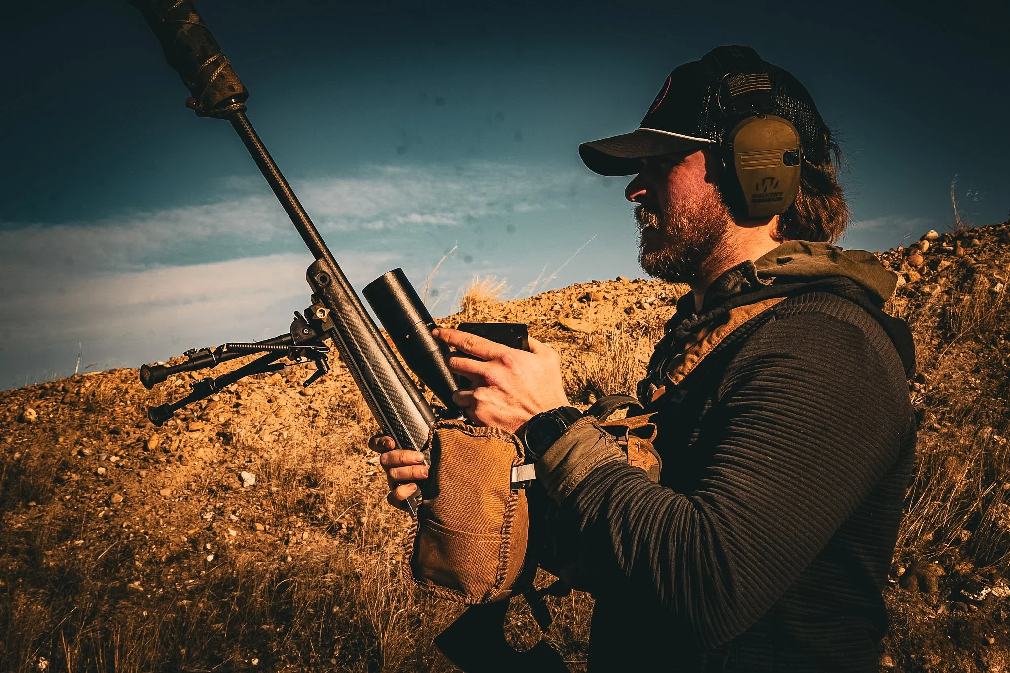 A person wearing headphones, a cap, and outdoor clothing, holding a rifle with a scope in a desert landscape.