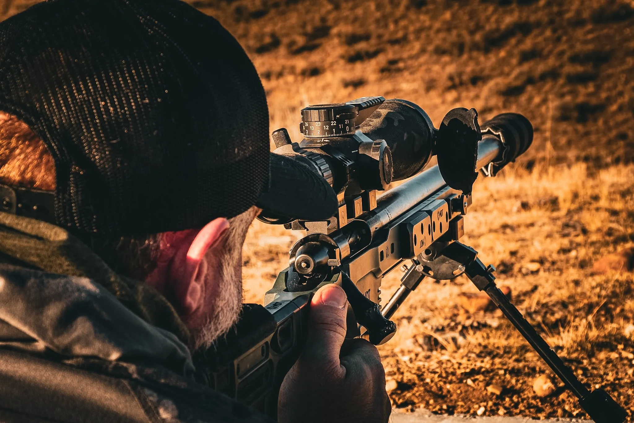 A person aiming a scoped rifle outdoors on a sunny day, with dry grass on the ground.