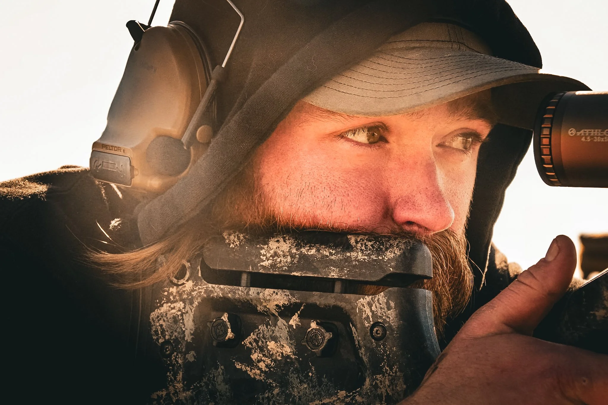 Close-up of a man wearing a hood and baseball cap, looking through a rifle scope with a serious expression, outdoors during sunset.
