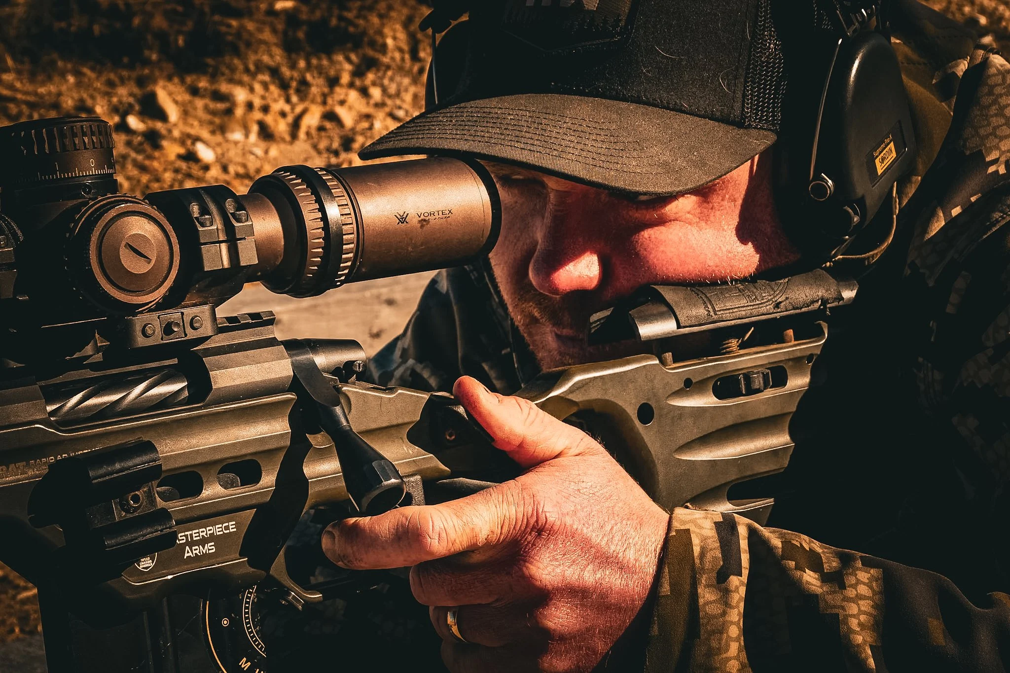 A person looking through a riflescope while aiming a rifle, wearing camouflage clothing, a tan baseball cap, and earp protection, outdoors on a sunny day.