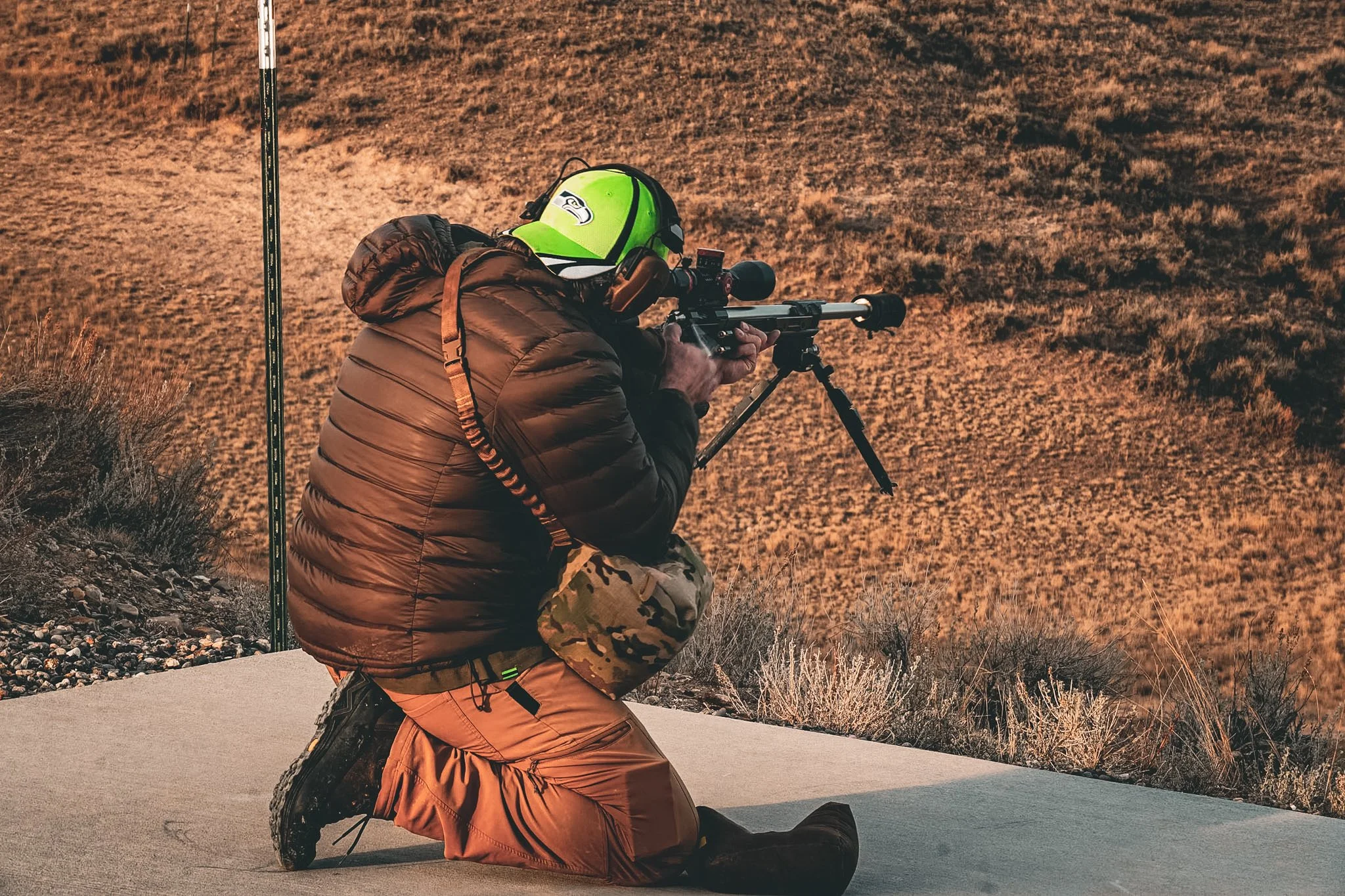 A person kneeling on one knee on the ground, aiming a rifle with a scope, in an outdoor setting with dry, shrub-covered terrain in the background, wearing a bright green helmet and camouflage clothing.