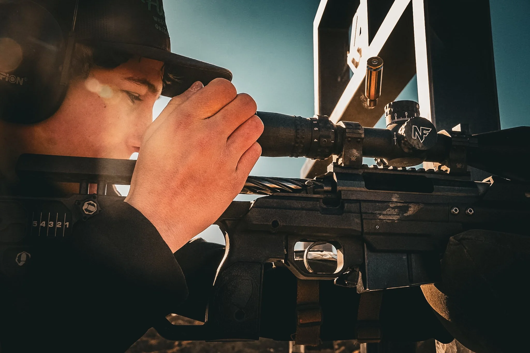 A person aiming a sniper rifle outdoors, wearing a hat and headphones, with a clear sky in the background.