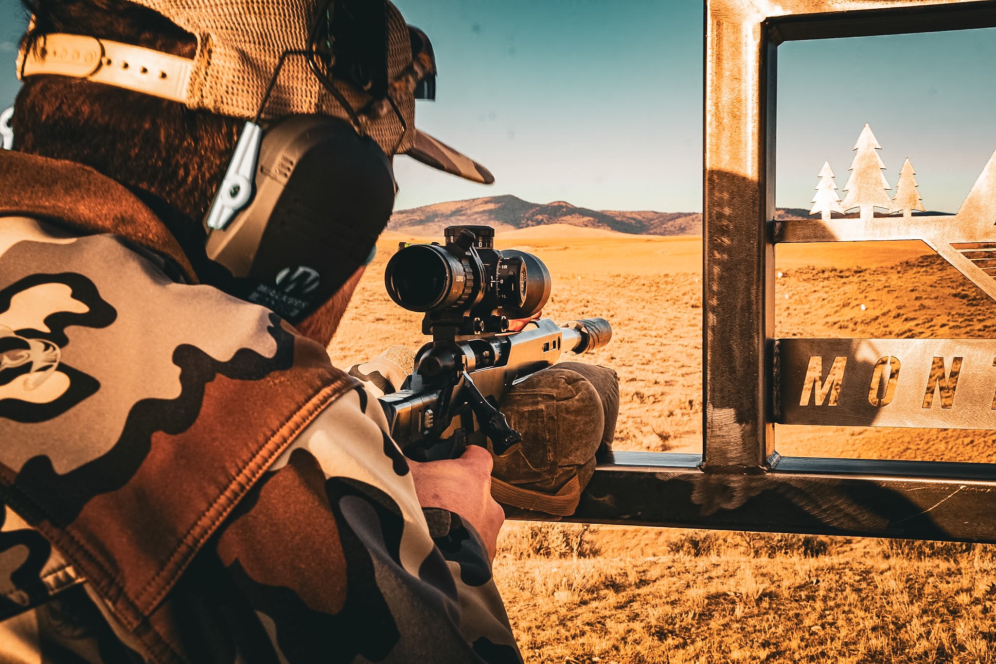 A person in camouflage clothing and a hat is aiming a scoped rifle from a mounted position, looking out onto a dry, open field with mountains in the distance during sunset.