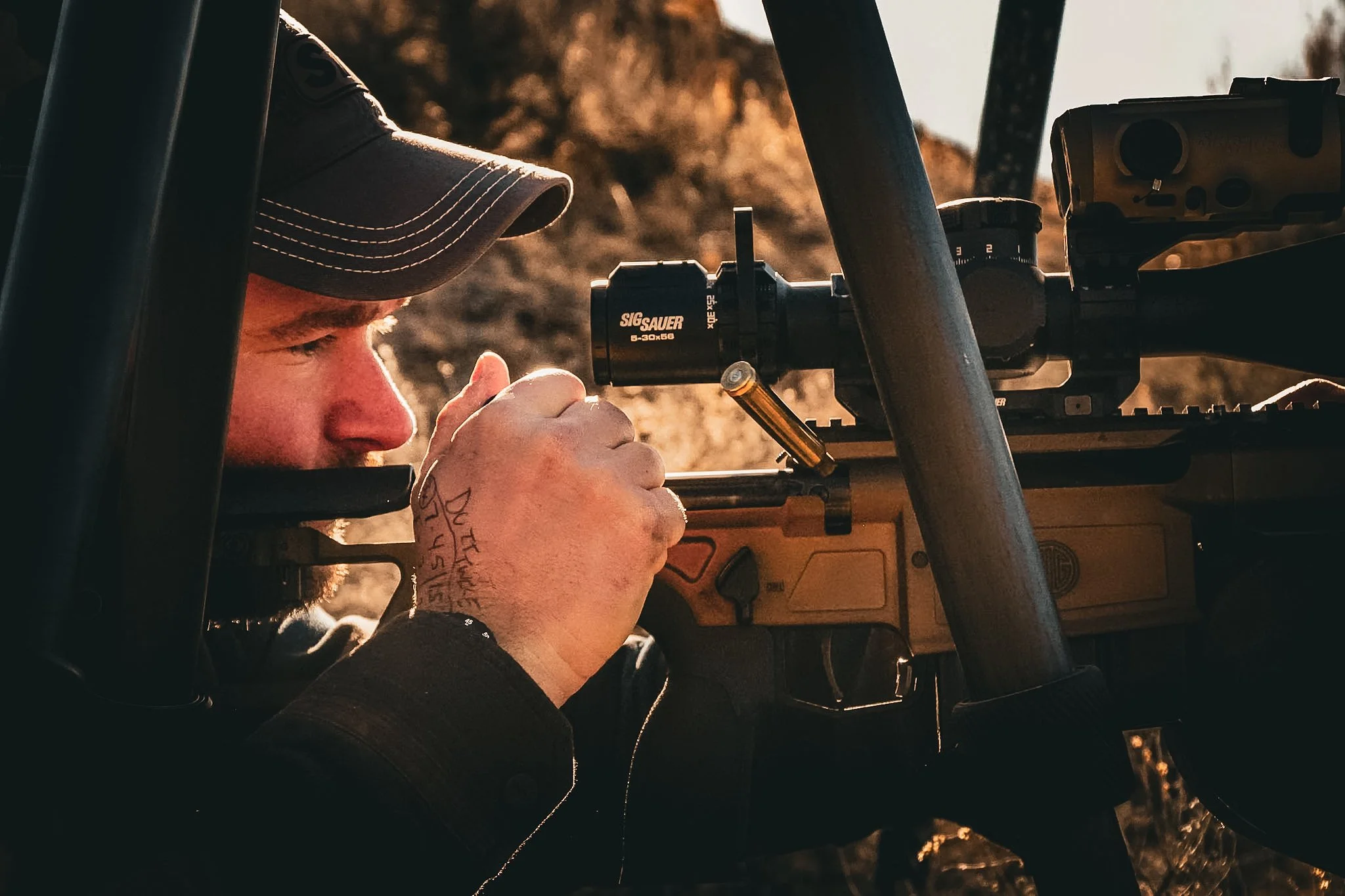 A person wearing a baseball cap and a dark jacket, adjusting the scope of a rifle in an outdoor setting during sunset.