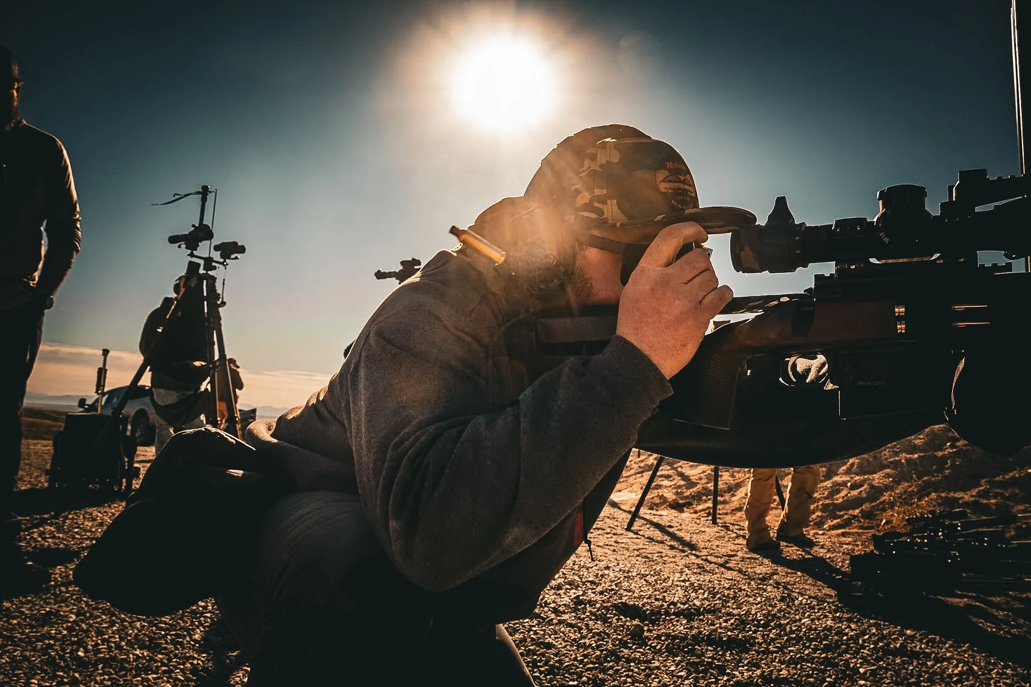 A military sniper is lying on the ground aiming through a scoped rifle with the sun setting behind him. There are other people and equipment in the background on a rough outdoor terrain.