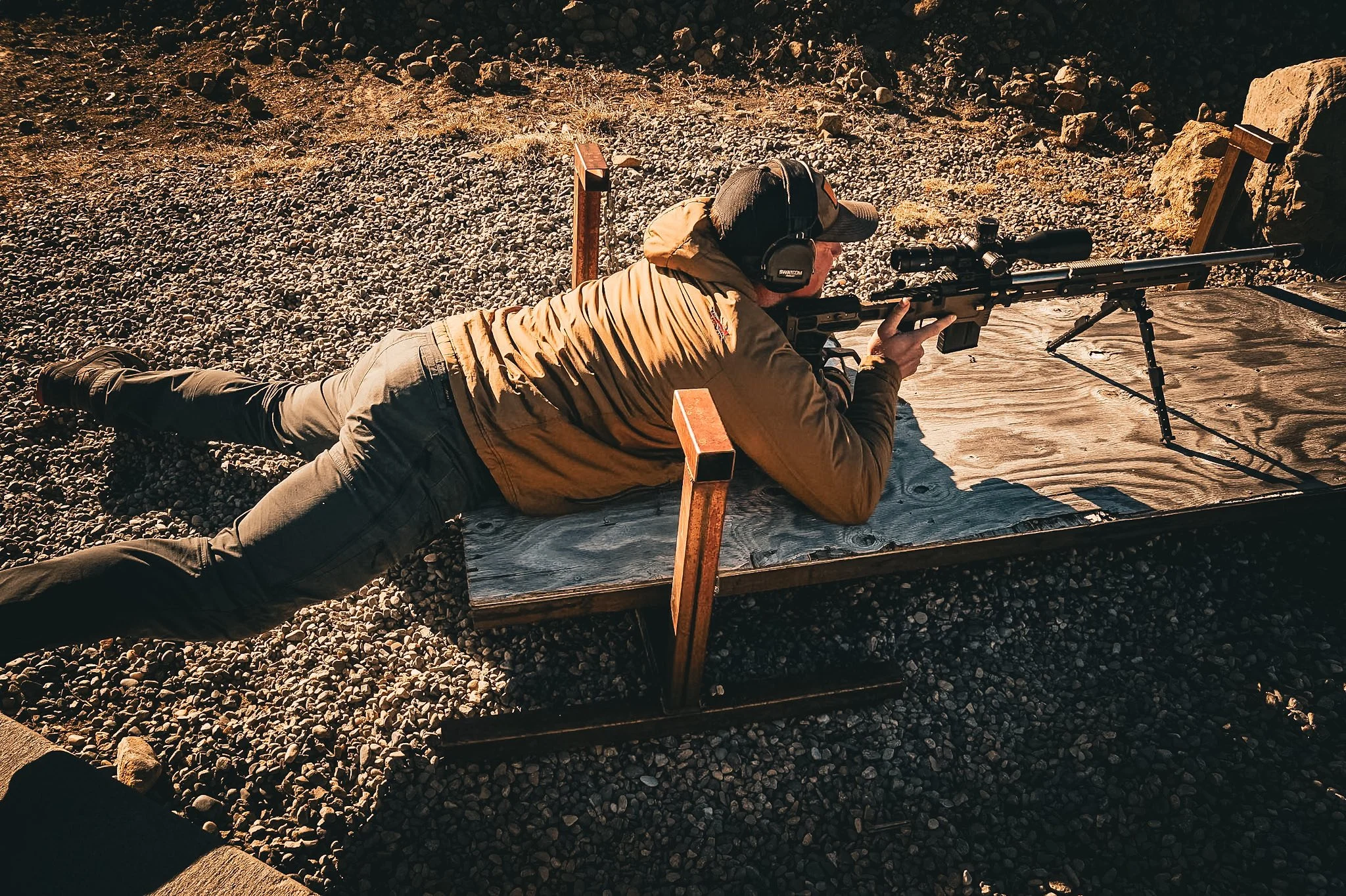 A man lying prone on a shooting platform, aiming a scoped rifle on a firing range.