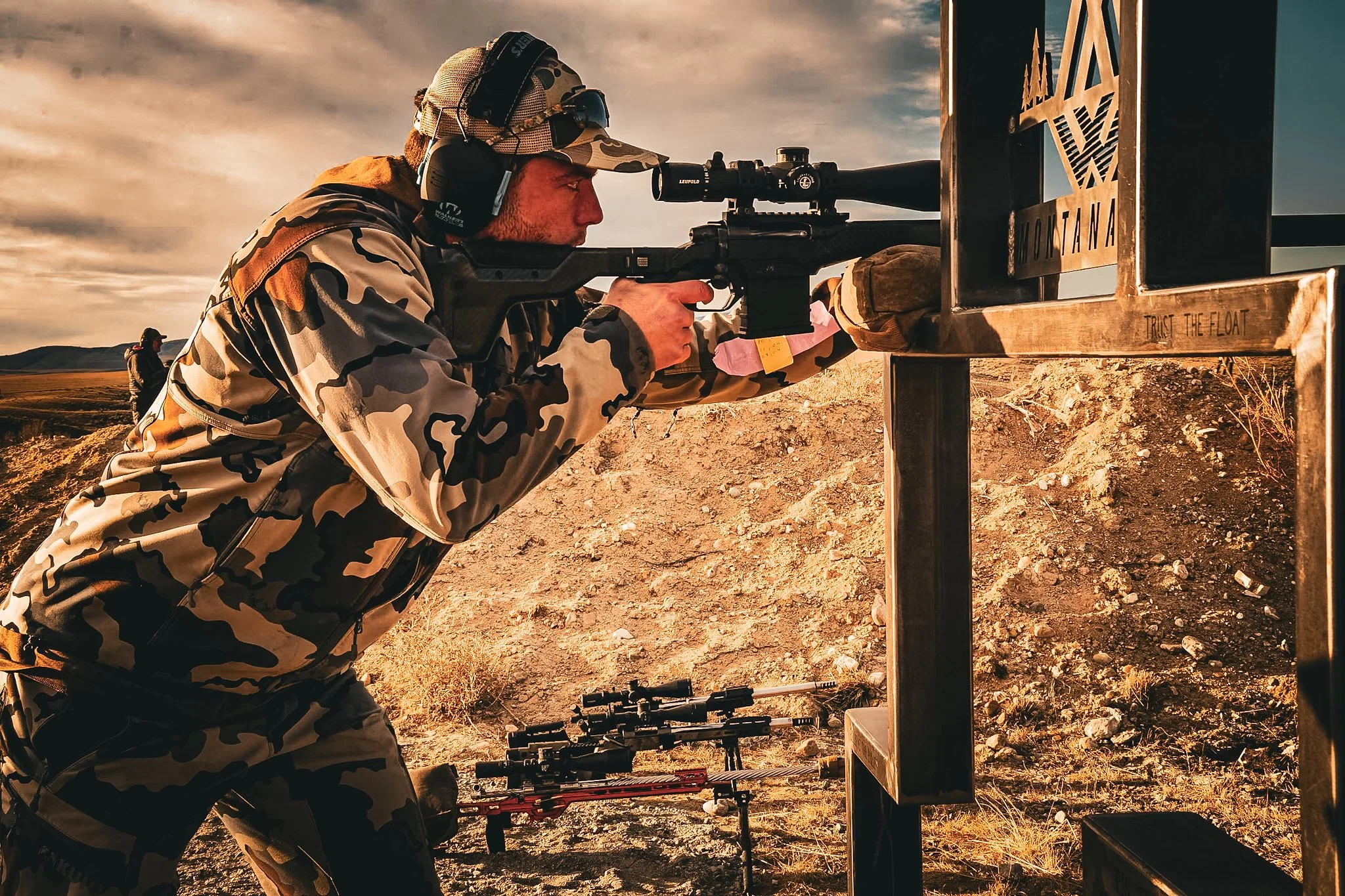 Person in camouflage gear aiming a scoped rifle at a shooting range, with other rifles on the ground nearby and a sunset sky in the background.
