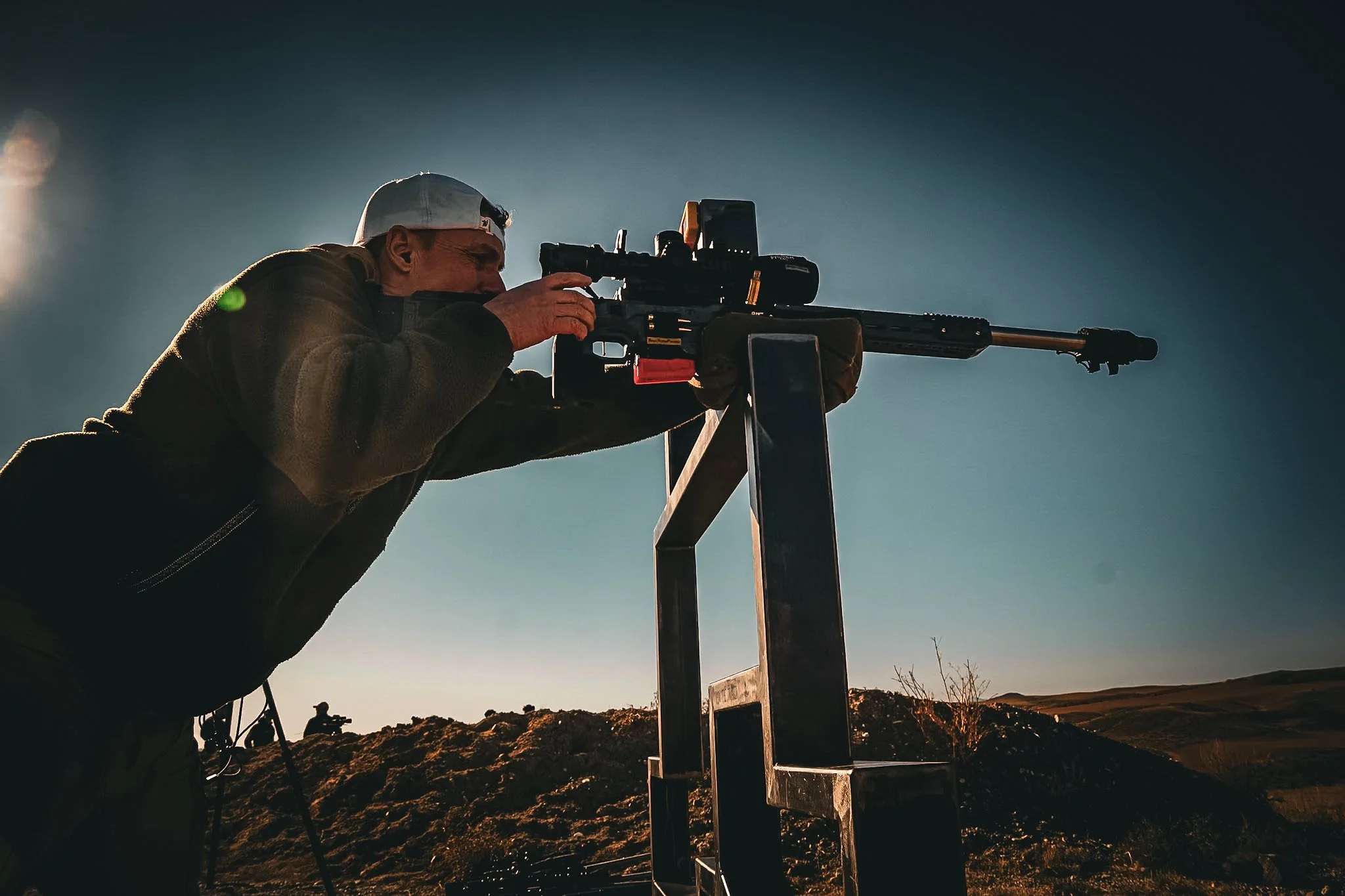 A man wearing a gray cap and dark jacket aims a sniper rifle on a tripod, outdoors during sunset or sunrise, with a hilly landscape in the background.