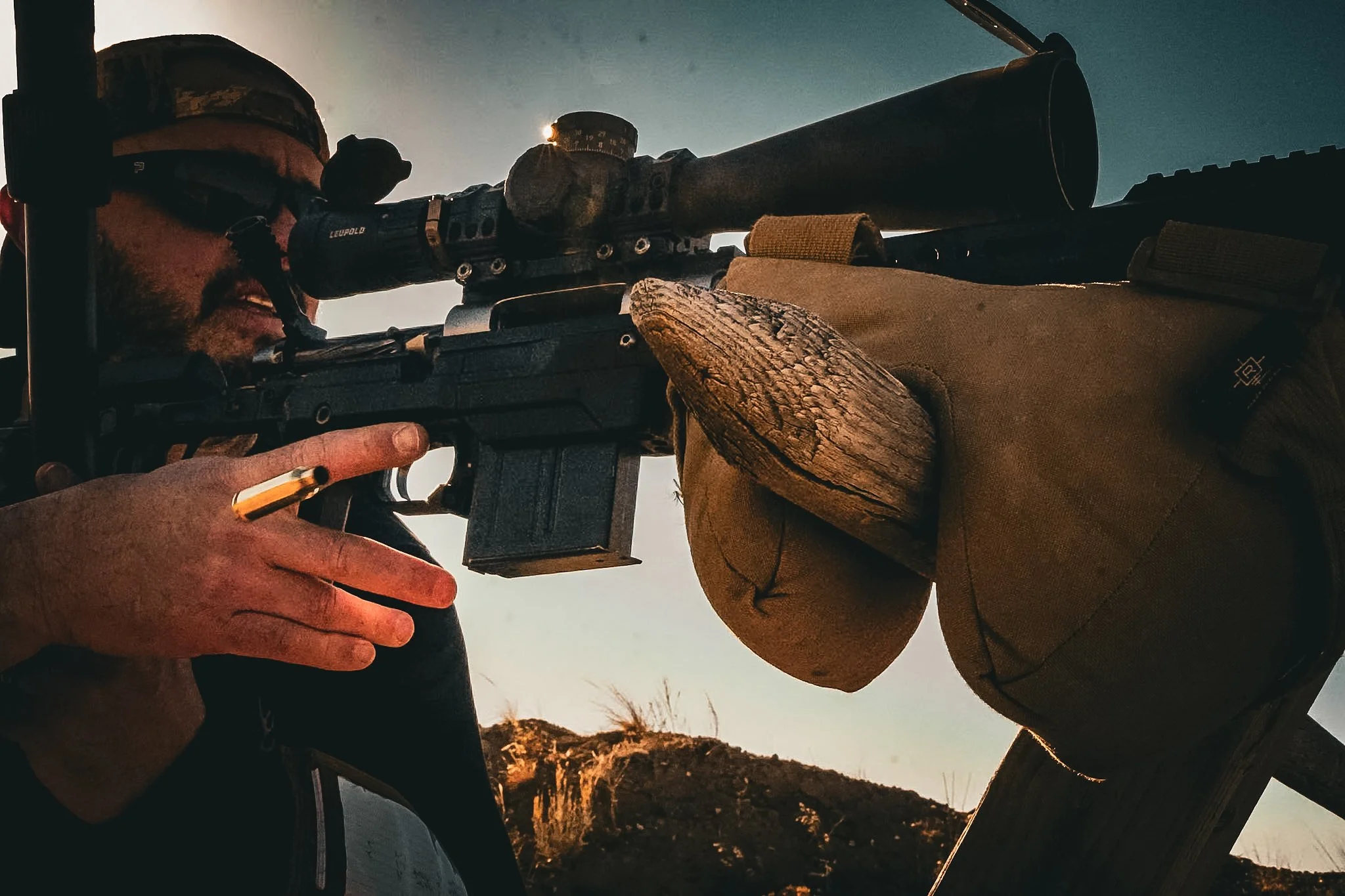 A man wearing glasses, a cap, and gloves looking through a scoped rifle outdoors at sunset or sunrise.