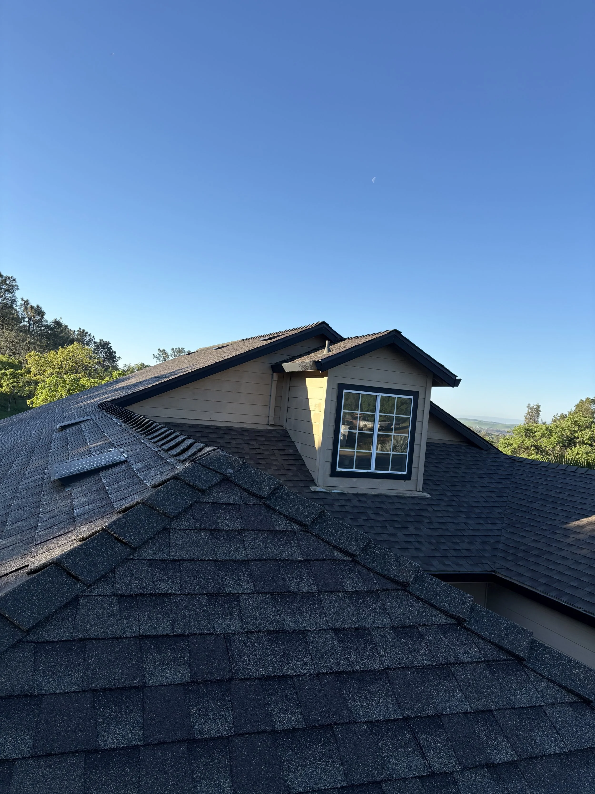 A residential house rooftop with asphalt shingles, and a dormer window under a clear blue sky with a visible crescent moon.