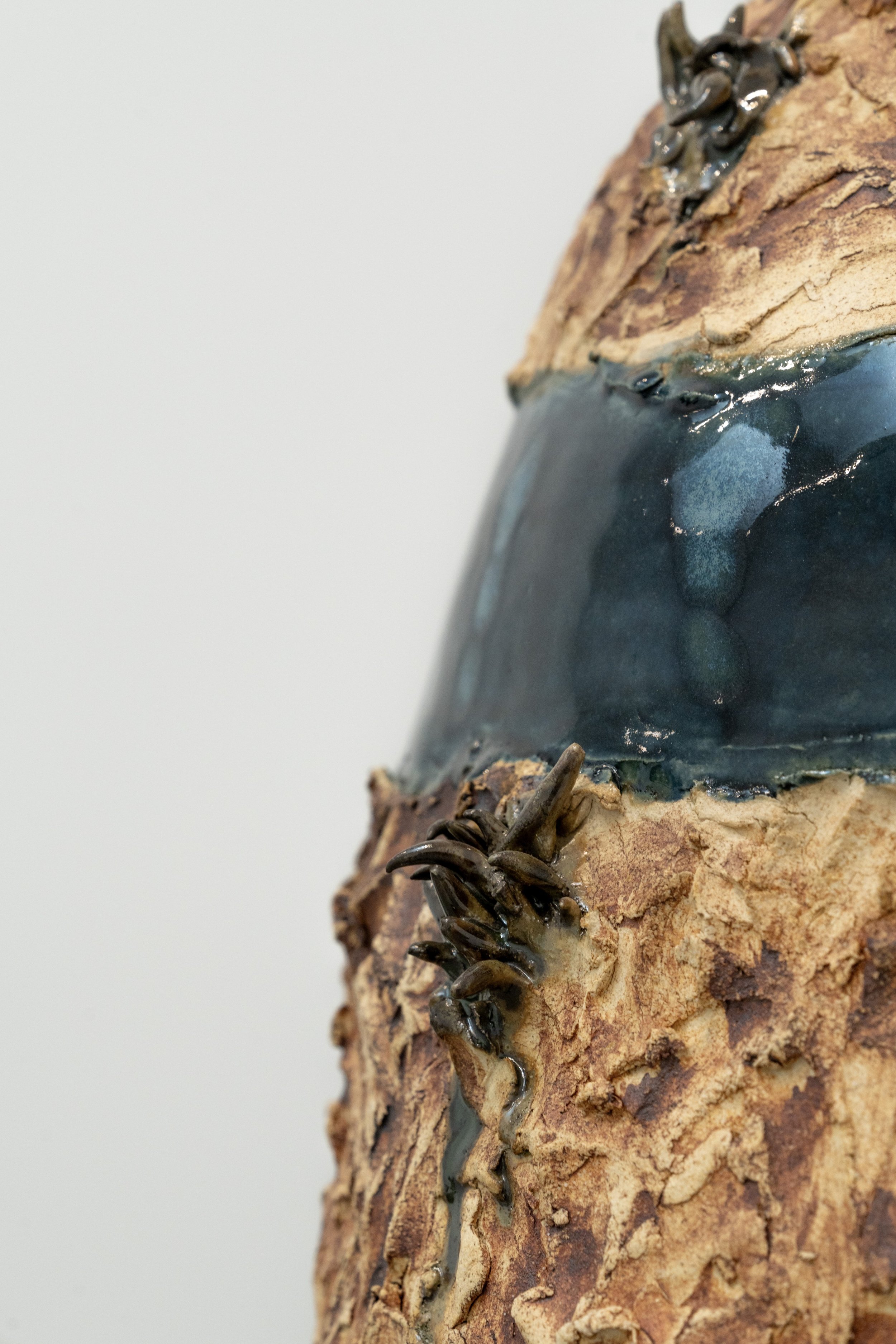 Close-up of a textured ceramic or clay sculpture with rough, bark-like surface, dark glazed areas, and small metal details resembling claws or talons.