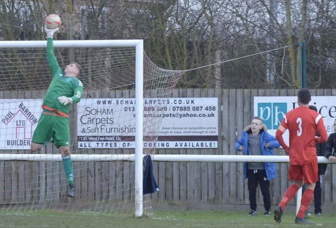 Soccer goalkeeper in green uniform catching a soccer ball in the goal during a match, with two players and a woman watching nearby.