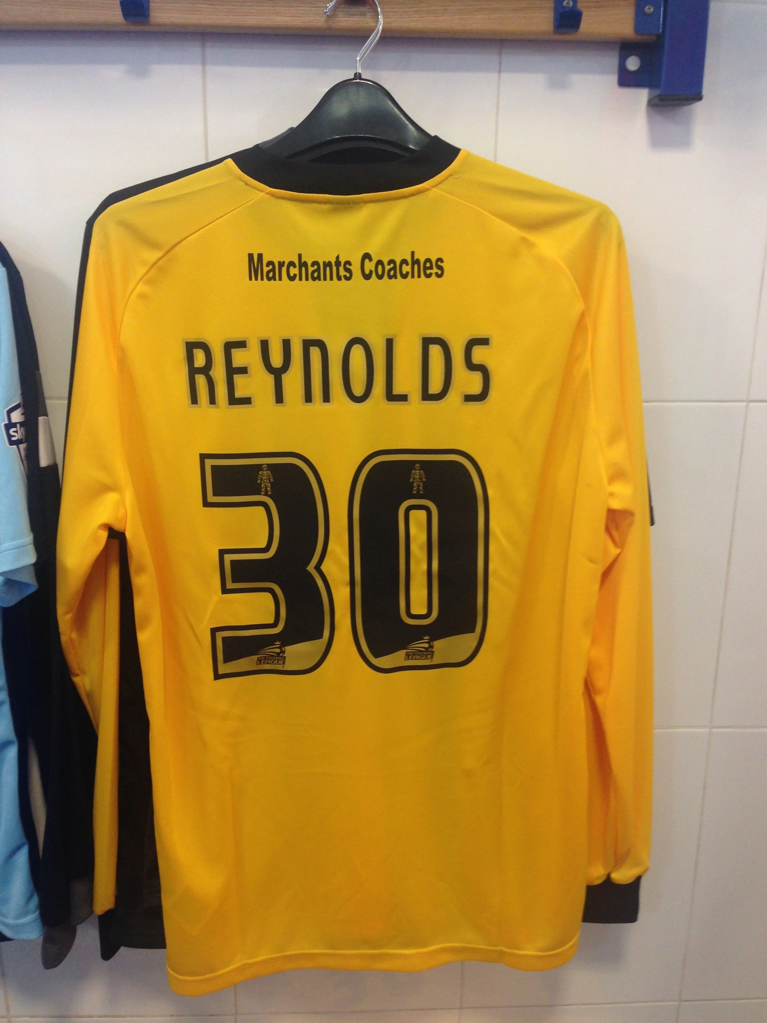 Yellow sports jersey hanging on a black hanger, with 'Reynolds' and the number '30' printed on the back. The jersey also has the text 'Marchants Coaches' at the top.
