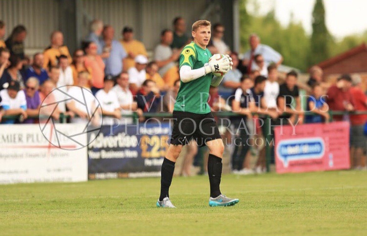 Soccer goalkeeper in green jersey and black shorts holding a soccer ball during a game, with spectators in the background.