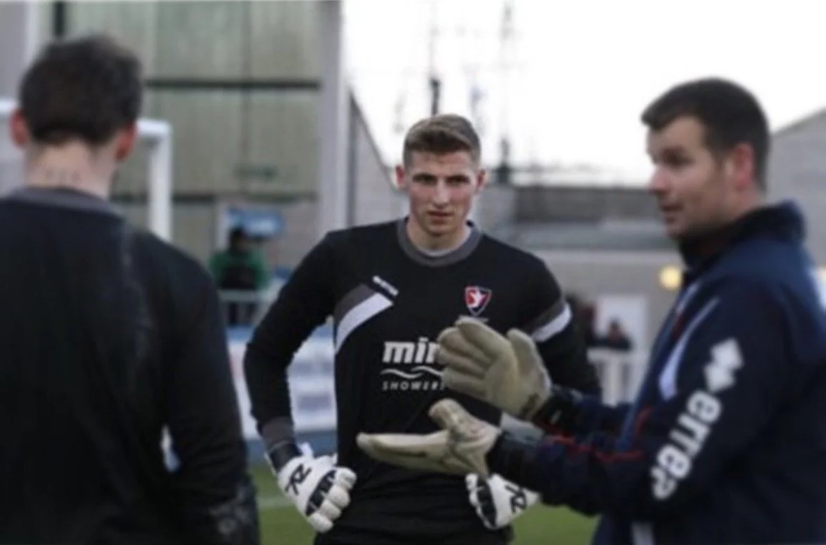 Three men, one in black sports gear, including gloves, standing outdoors on a sports field, engaged in conversation.